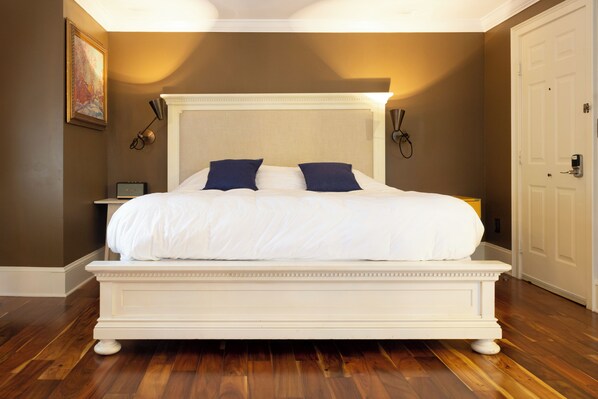 A large bed with white bedding and two dark pillows, a nightstand with a lamp, and a painting on the wall.