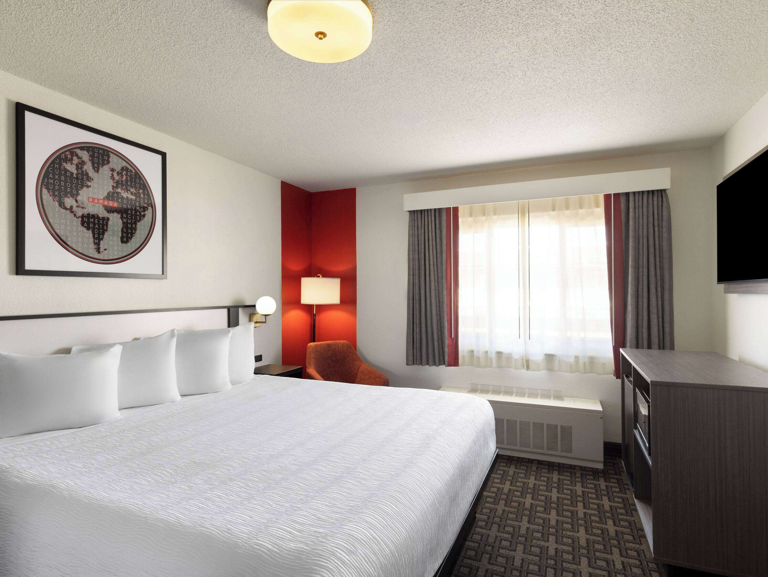 A hotel room with a large bed, a desk with a flat-screen TV, a red accent wall, and a window with curtains.