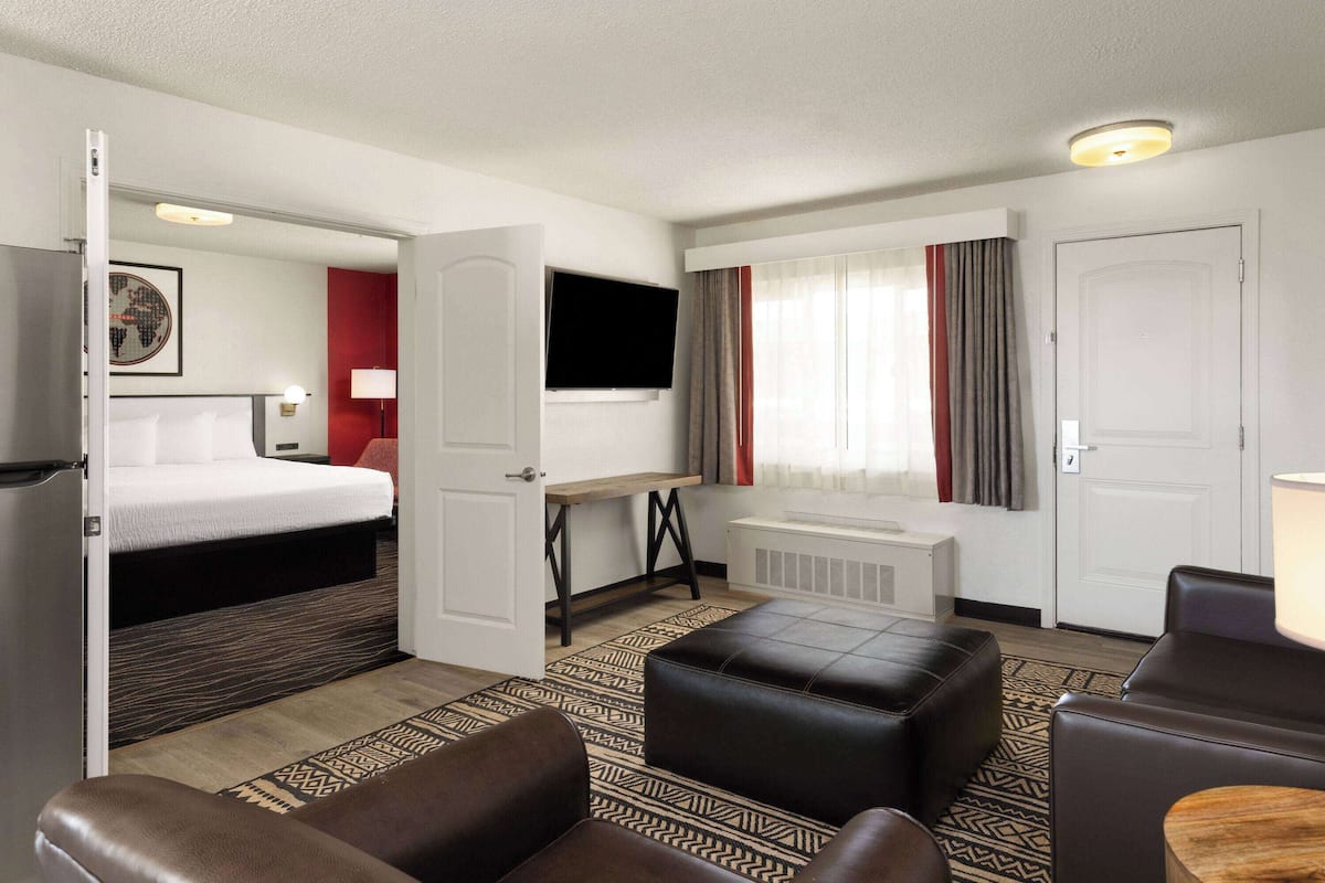 A hotel room with a bed, a desk, a TV, a seating area with ottomans, and a window with curtains.
