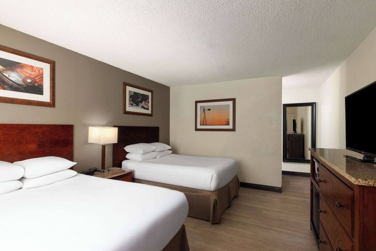 A hotel room with two beds, a desk, a lamp, a TV, and artwork on the walls.