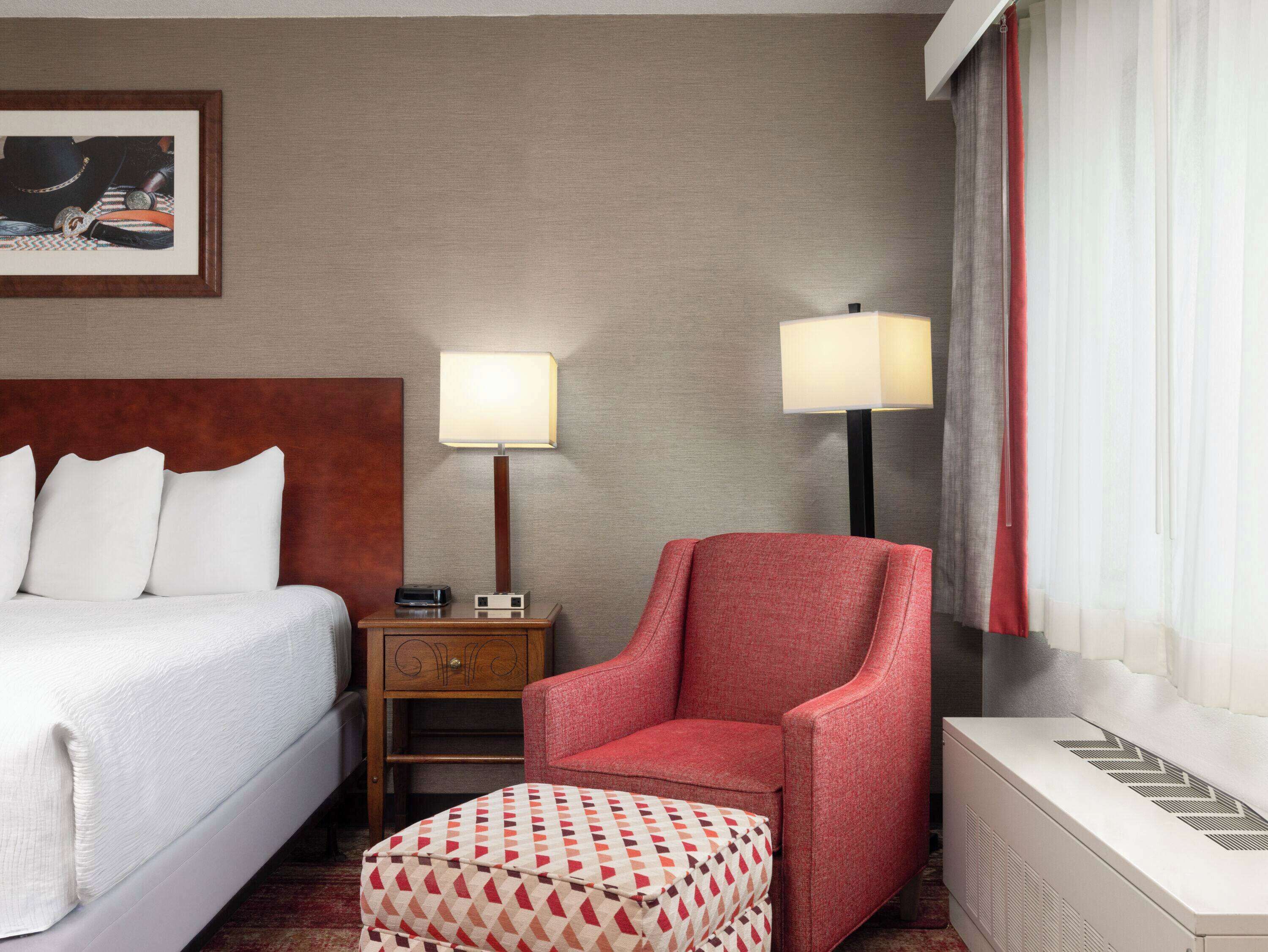 A hotel room with a bed, a red armchair, a patterned ottoman, a nightstand with a lamp, and a radiator.