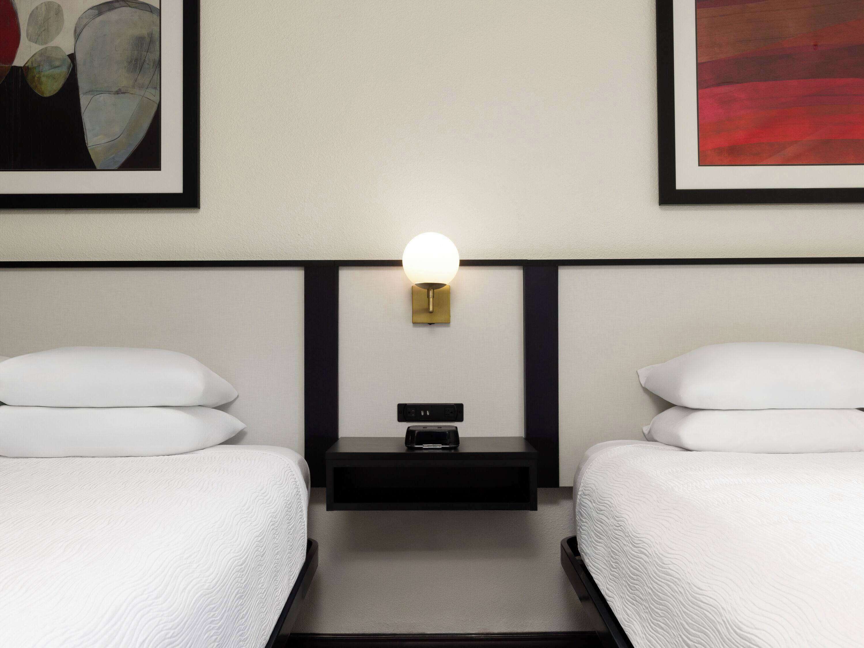 A hotel room with two beds, a nightstand with a lamp, and artwork on the walls.