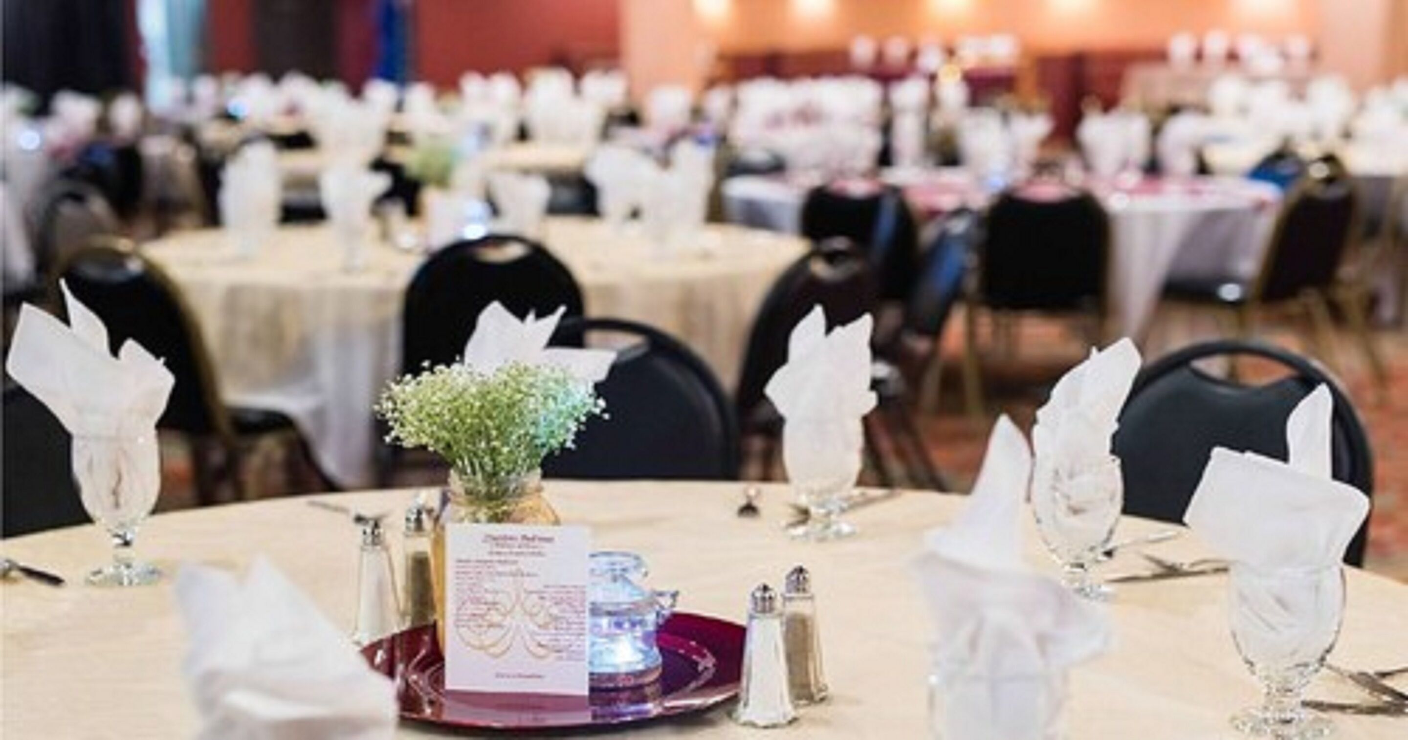 A banquet hall with round tables set for a formal event, featuring white napkins, glassware, and a centerpiece with flowers.