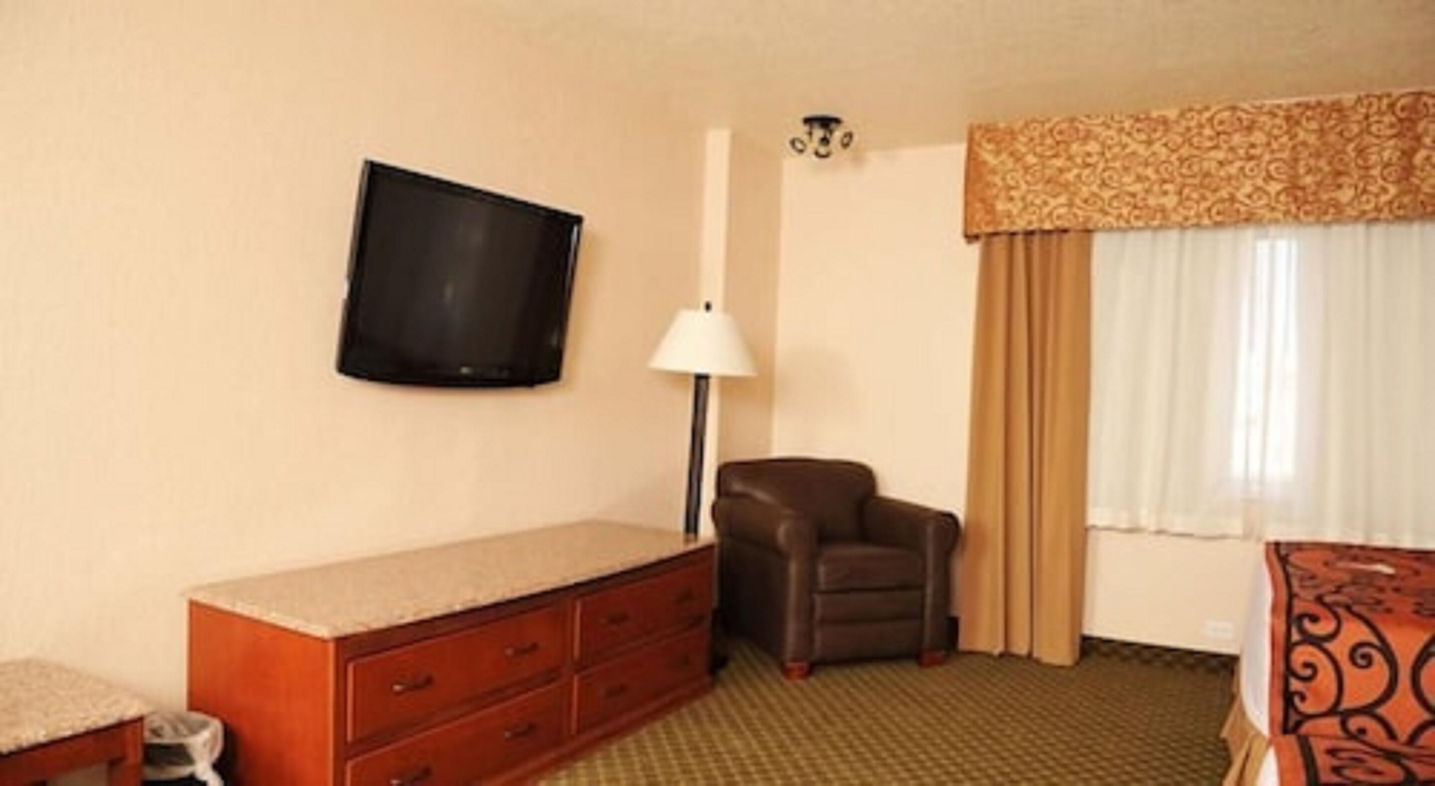Une chambre d’hôtel dotée d’un téléviseur à écran plat fixé au mur, d’un fauteuil en cuir, d’une commode en bois avec des tiroirs, d’un lampadaire et d’un lit recouvert d’une couvre-lit à motifs.