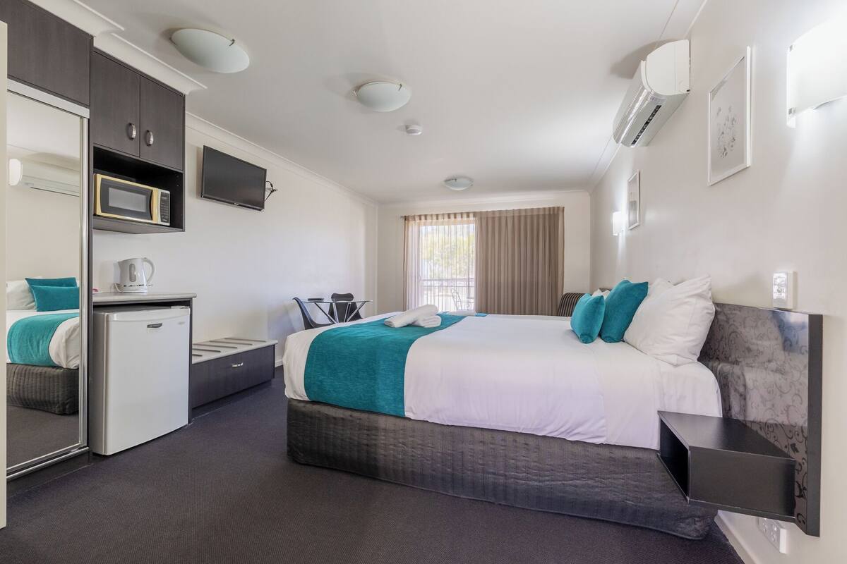 A modern hotel room with a large bed, a desk with a chair, a microwave, a coffee maker, and a TV.