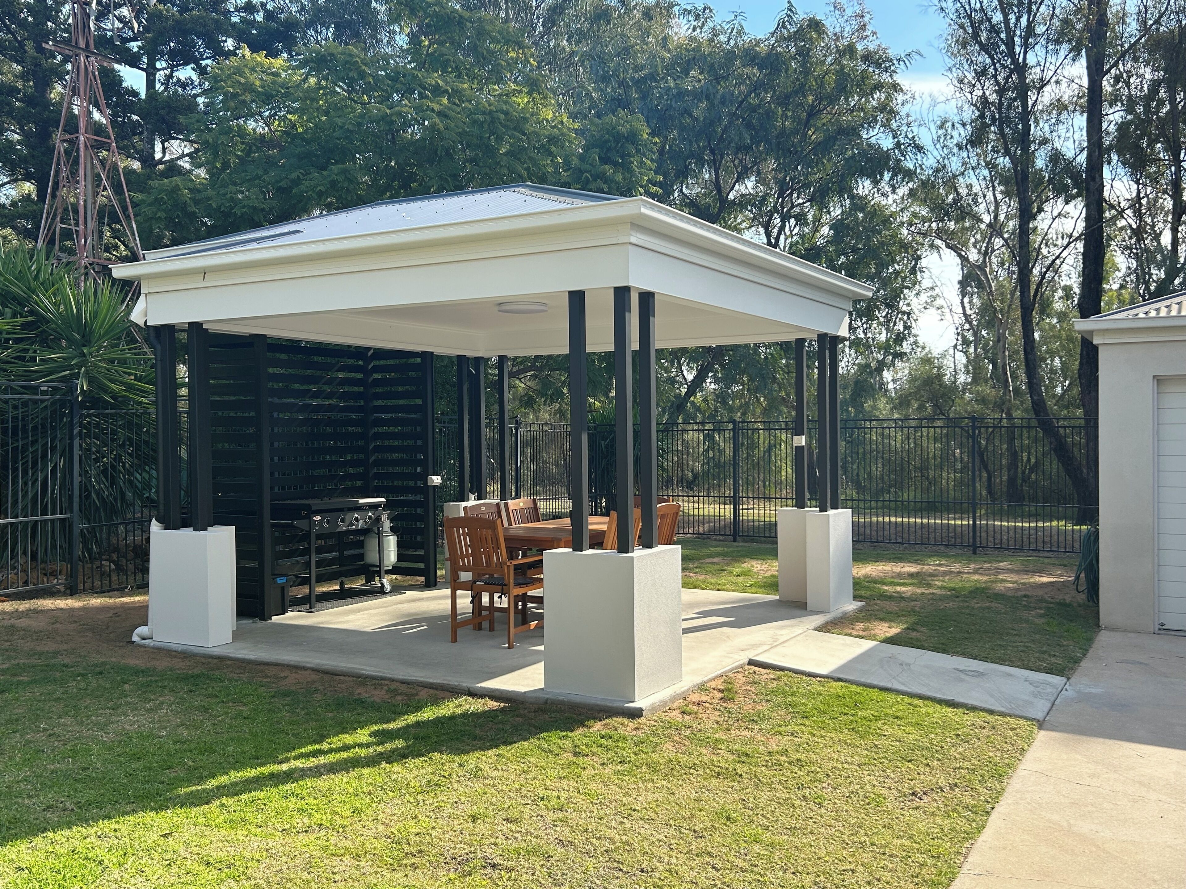 A covered patio area with a table and chairs, surrounded by a fence and trees.