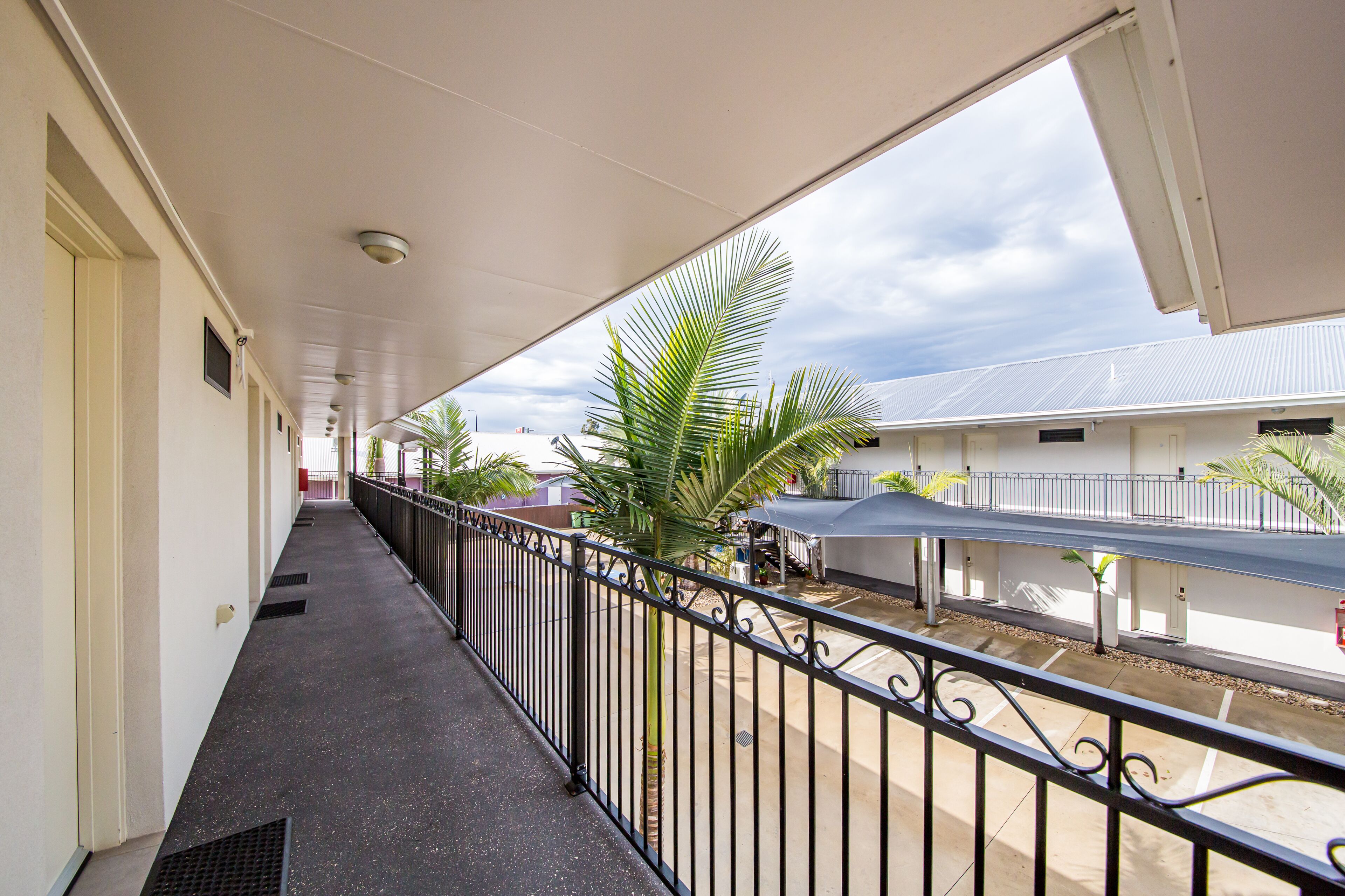 A covered corridor with a metal railing, palm trees, and a view of buildings in the background.