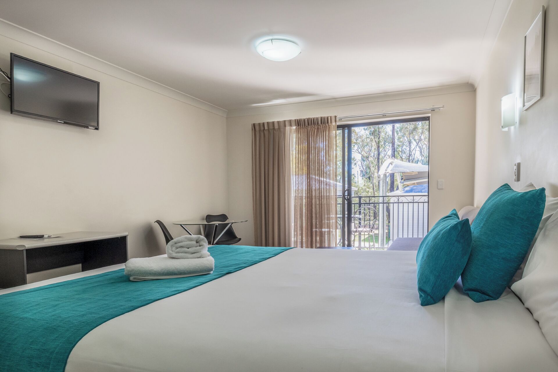 A hotel room with a bed, a TV mounted on the wall, a desk with a chair, a window with curtains, and a balcony with a railing.