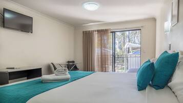 A hotel room with a bed, a TV mounted on the wall, a desk with a chair, a window with curtains, and a balcony with a railing.