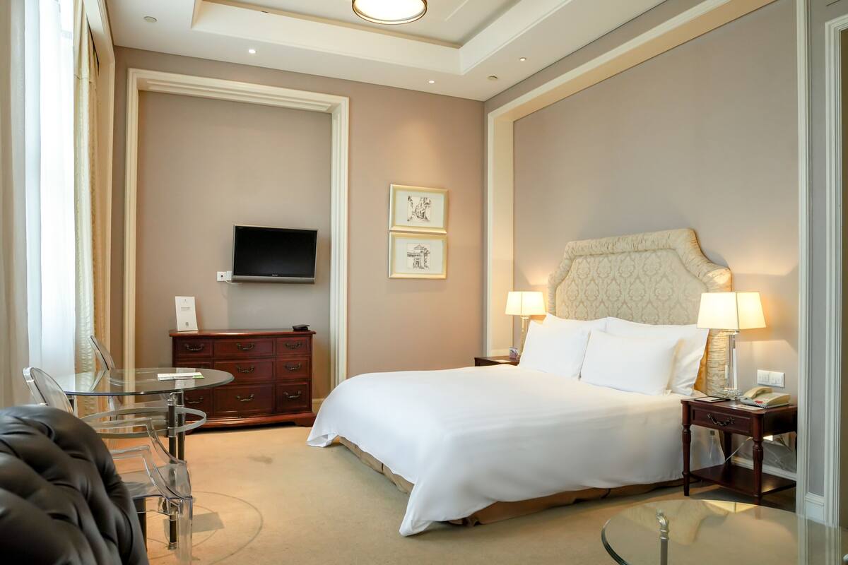 A hotel room with a large bed, a TV mounted on the wall, a glass table, and a leather chair.