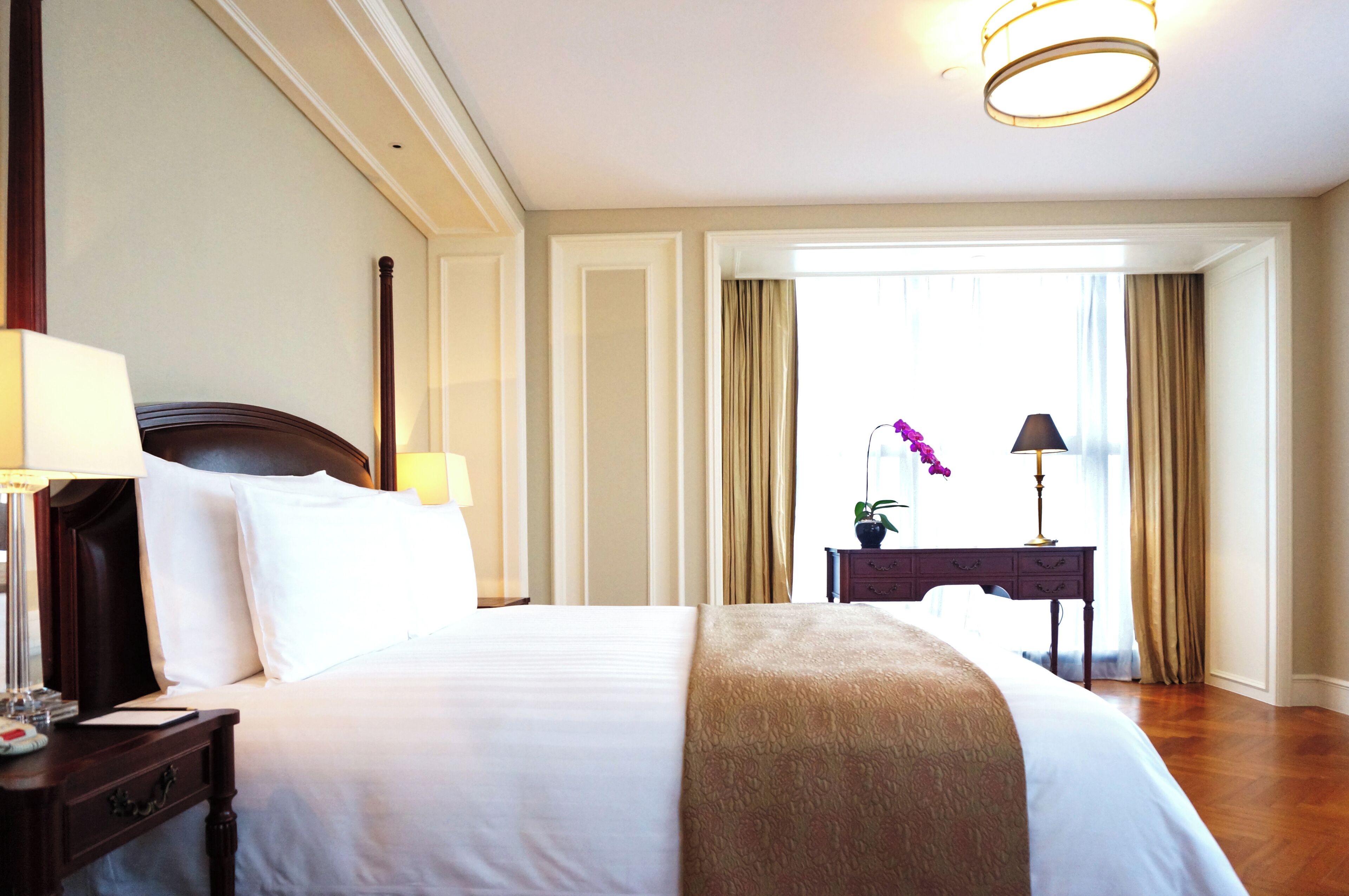 A hotel room with a large bed, a nightstand with a lamp, a dresser, and a view of the outdoors through a window with curtains.