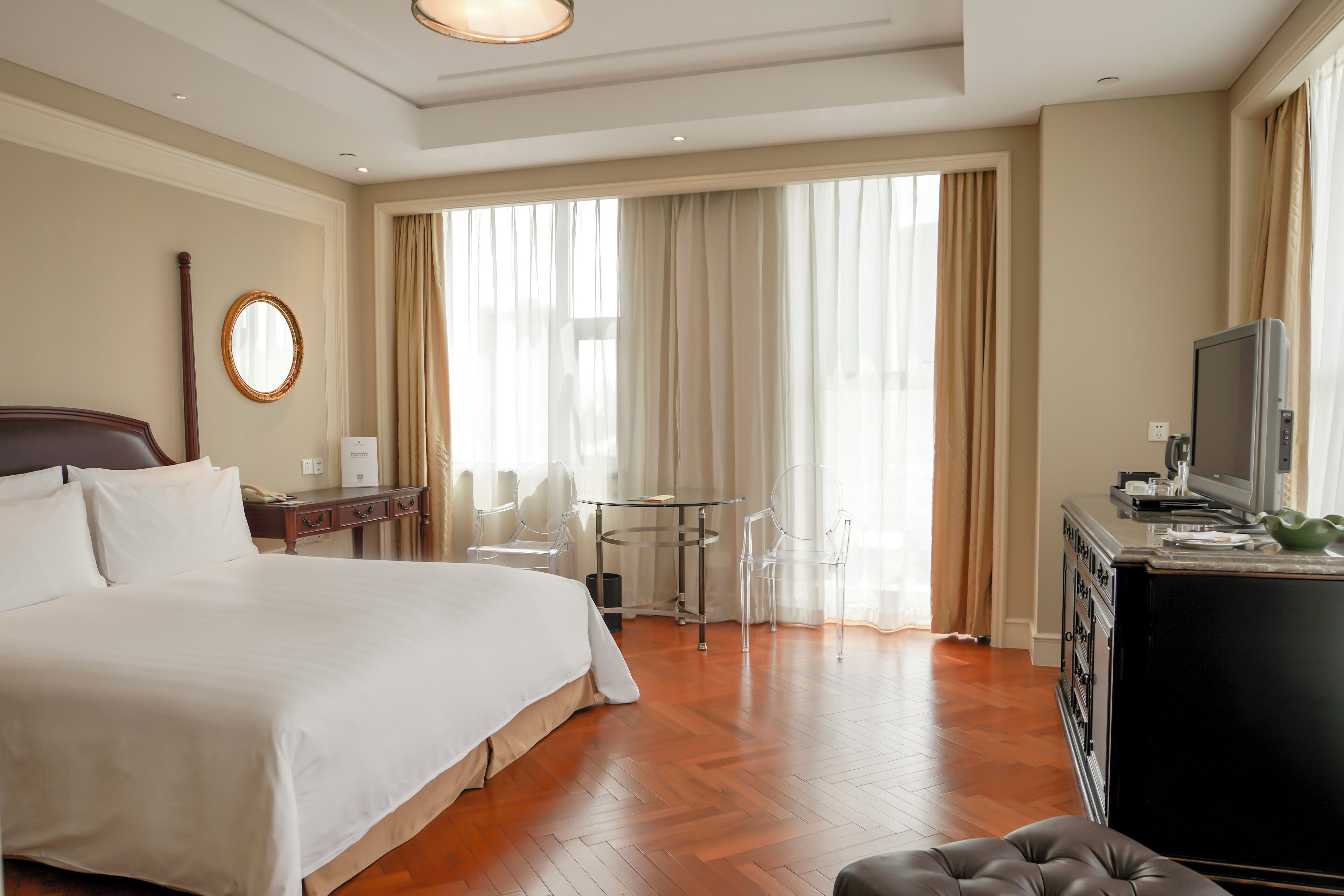 A hotel room with a large bed, a desk with a television, and a view of the outdoors through large windows.