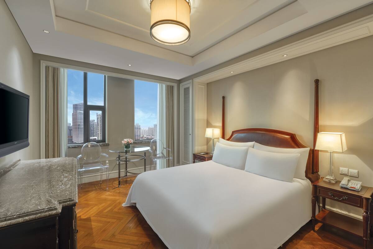 A hotel room with a large bed, a flat-screen TV, a desk with a chair, a small table with a chair, and a view of the city through large windows.