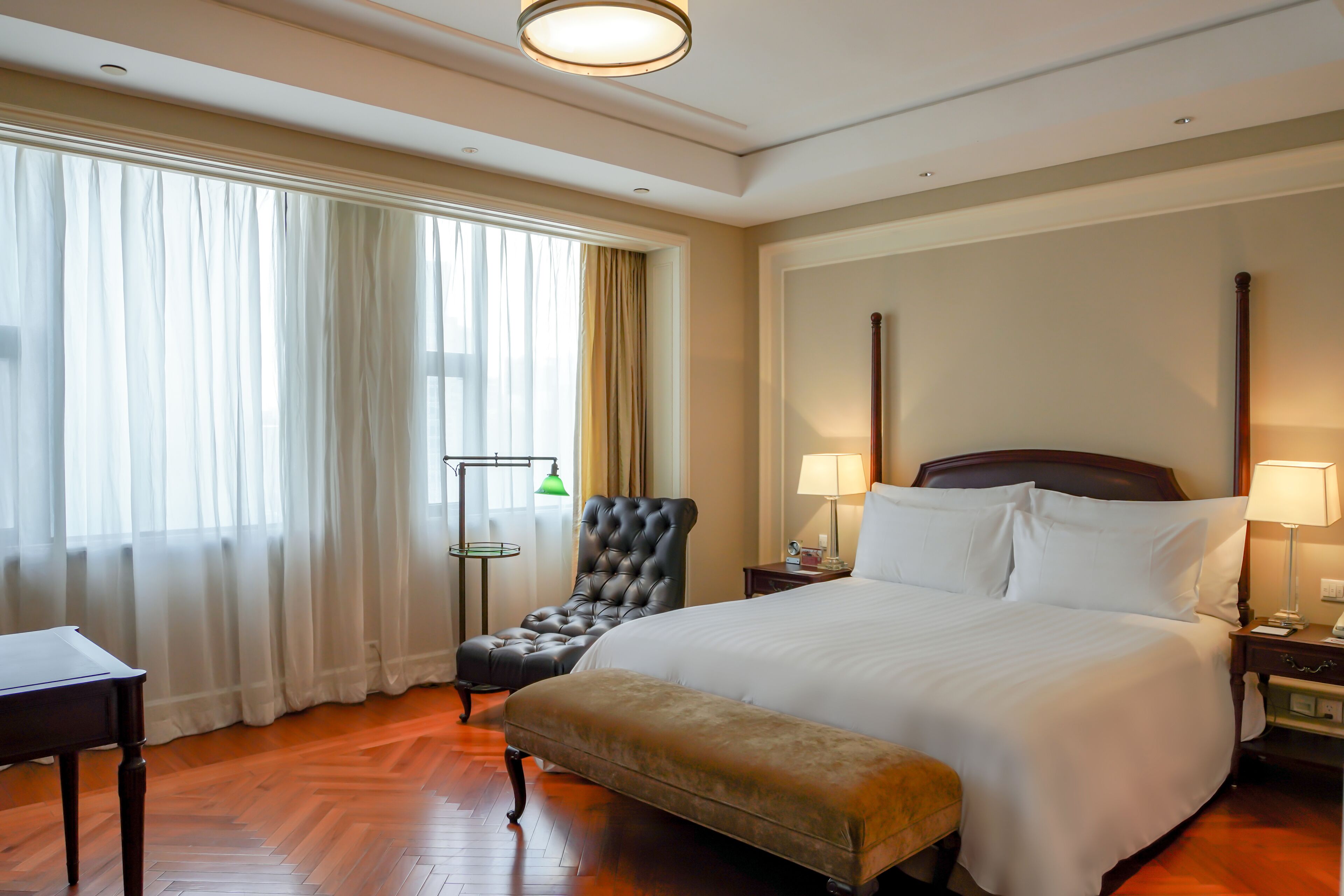 Ein Hotelzimmer mit einem großen Bett, einem Sessel, einer Bank, einem Nachttisch, einer Lampe und einem Fenster mit Vorhängen.