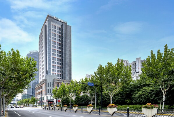 A tall hotel with a modern design, located on a city street with trees and a clear sky. - Sunrise on the Bund (Shanghai)