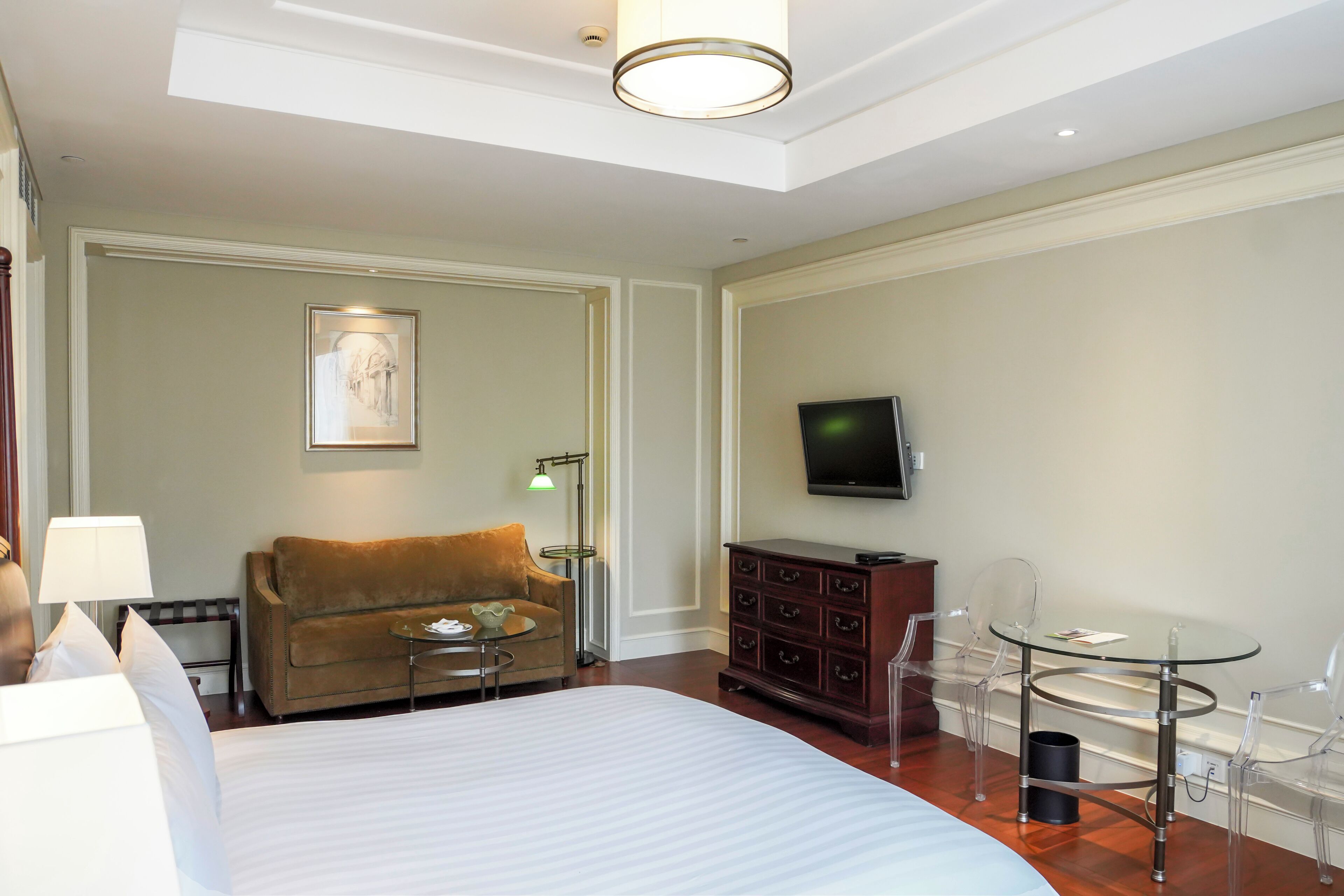 Una habitación de hotel con una cama grande, un sofá marrón, una pequeña mesa redonda, una mesa de centro de vidrio, un aparador de madera y un televisor montado en la pared.