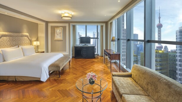 A hotel room with a large bed, a sofa, a coffee table with a vase, a TV, and a view of the city skyline. - Sunrise on the Bund (Shanghai)