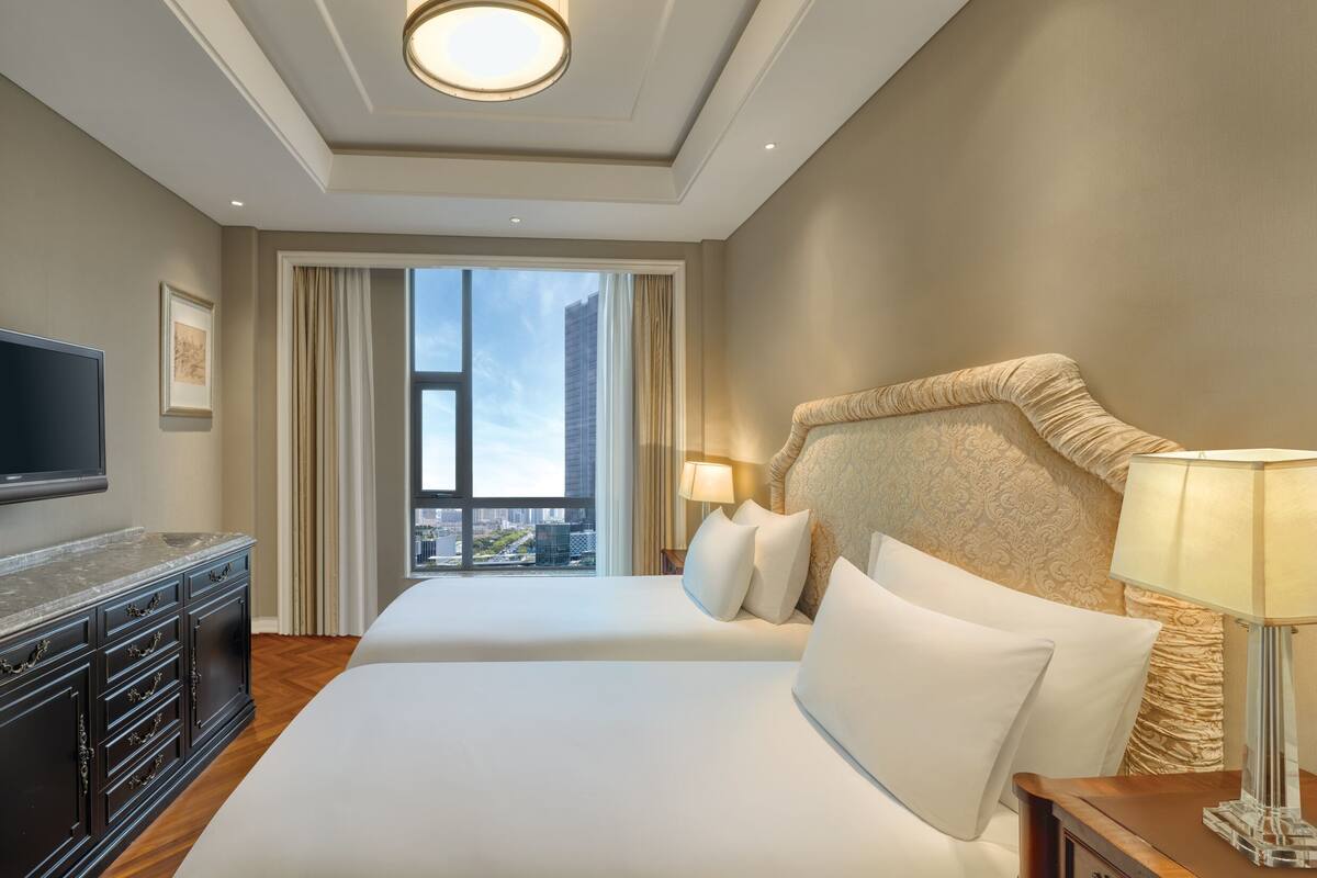A hotel room with a large bed, a TV, a dresser, and a view of the city through large windows.