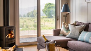 A cozy living room with a fireplace, a sofa with cushions, a table with a bottle and glasses, and a view through a glass door.