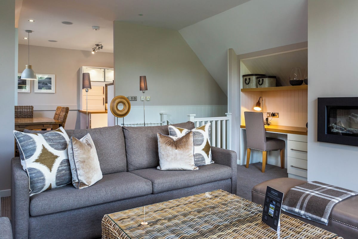A modern living room with a grey sofa, patterned cushions, a woven ottoman, and a dining area with a wooden table and chairs.