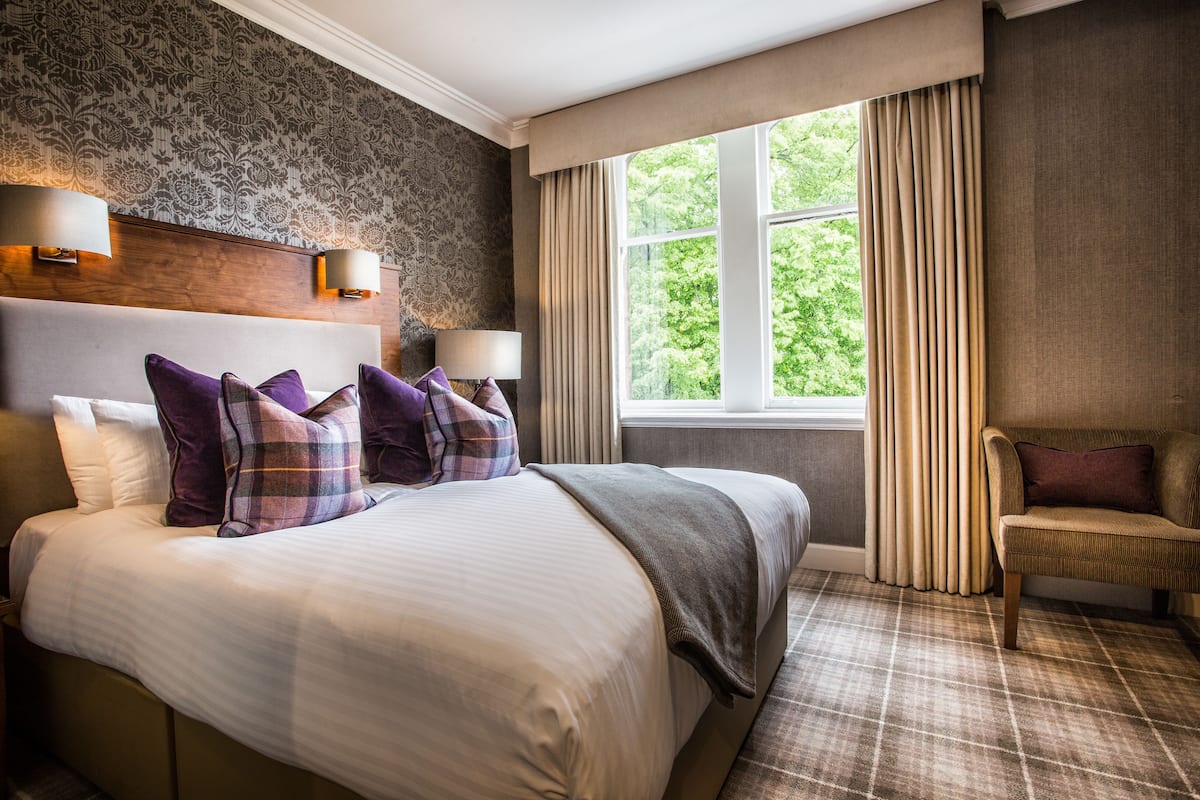 A hotel room with a large bed, two bedside lamps, a chair, a window with curtains, and a patterned wall.