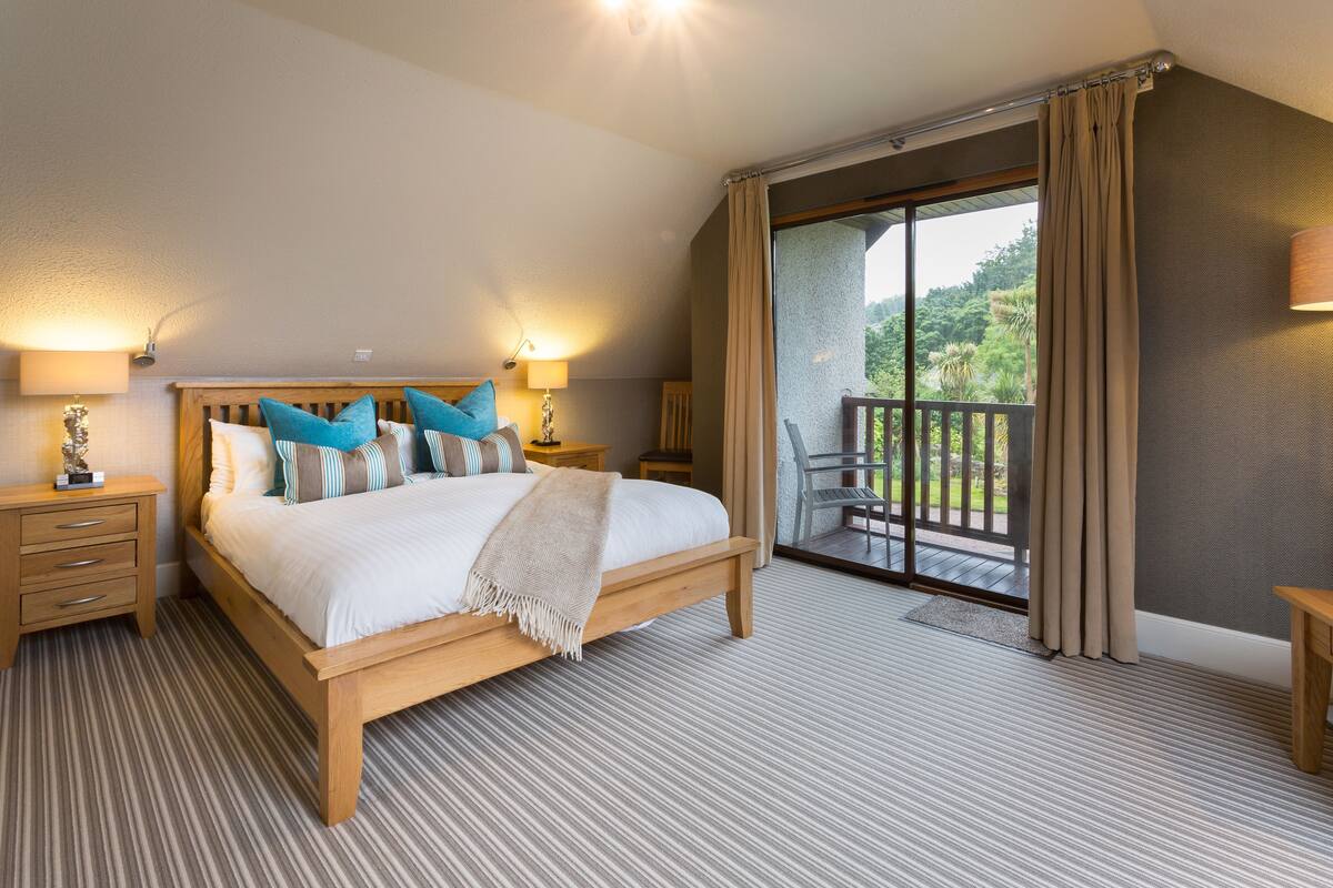 A bedroom with a large bed, wooden headboard, and two bedside tables with lamps. There is a balcony with a view of greenery.