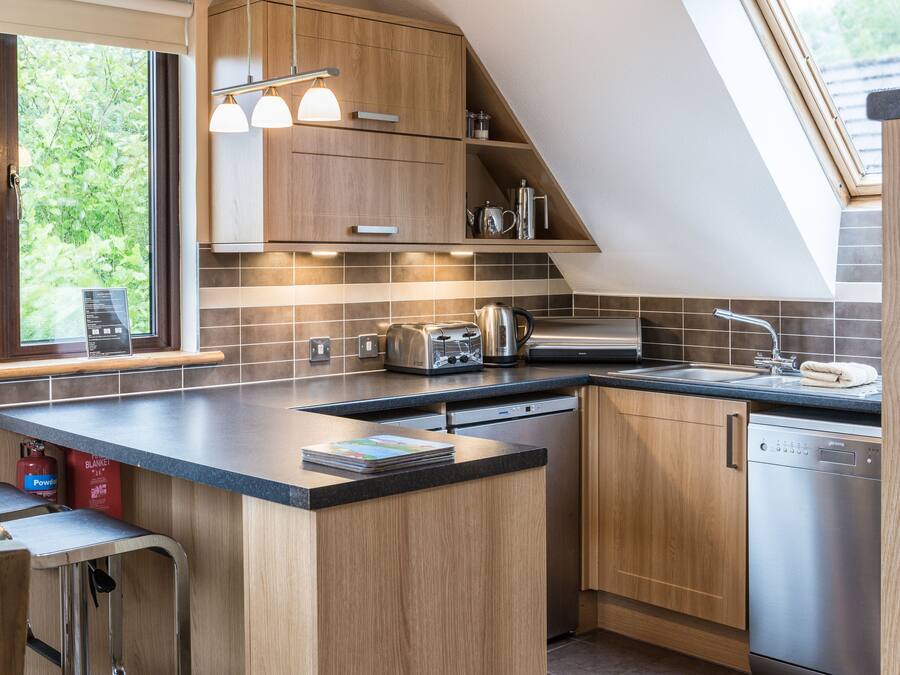 A modern kitchen with wooden cabinets, a black countertop, a built-in oven, a dishwasher, and a window with a view of greenery.