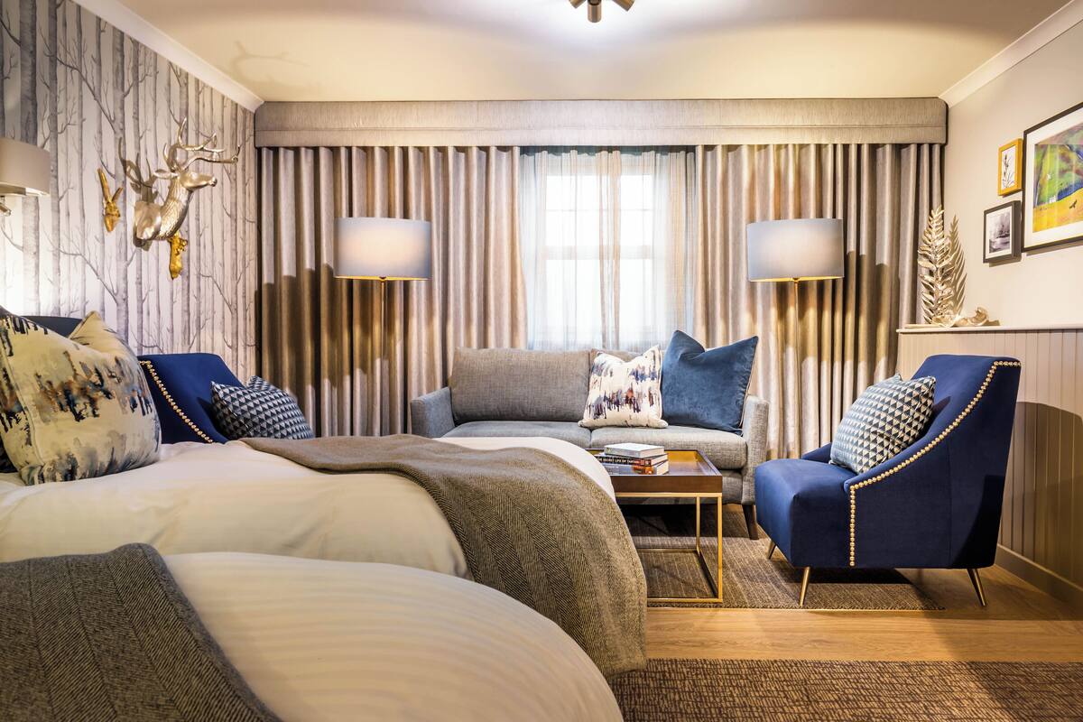 A hotel room with a large bed, a sofa, a chair, and a small table. There are curtains, a lamp, and wall art.