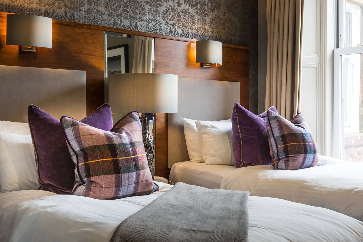 A hotel room with two beds, each with white linens and multiple pillows, including purple and plaid ones. There are wall-mounted lamps and a large mirror above the beds.