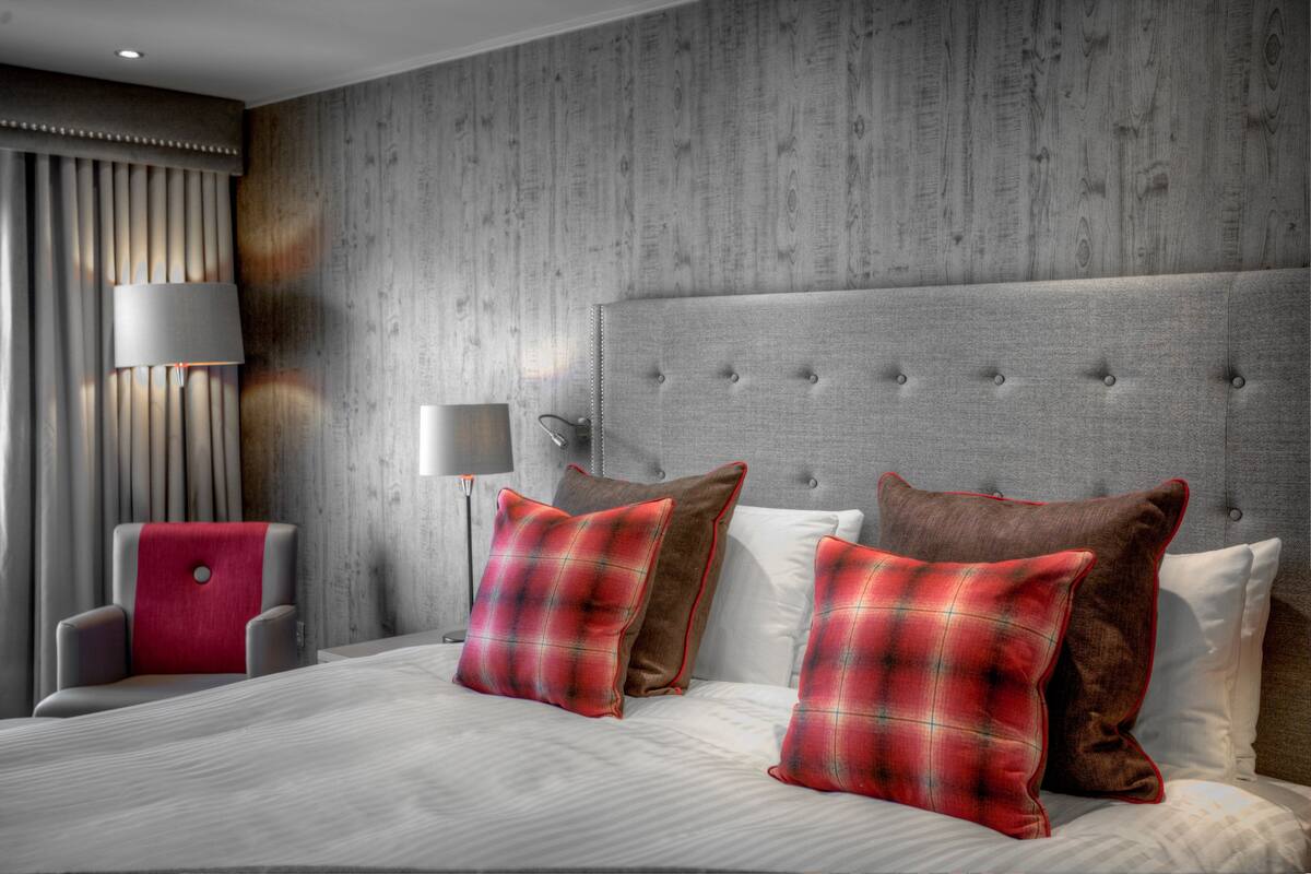 A hotel room with a large bed, grey upholstered headboard, and a red and brown plaid throw pillow.