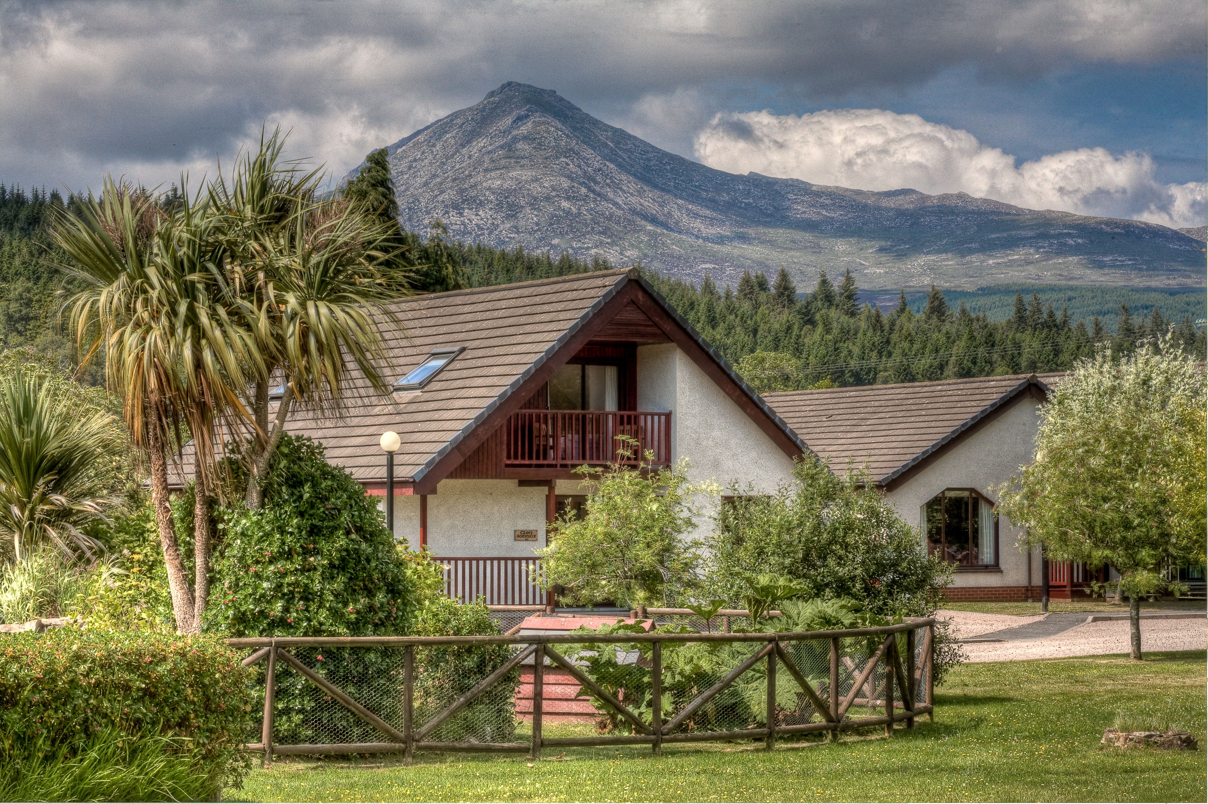 A two-story house with a balcony, surrounded by lush greenery and a mountain in the background.