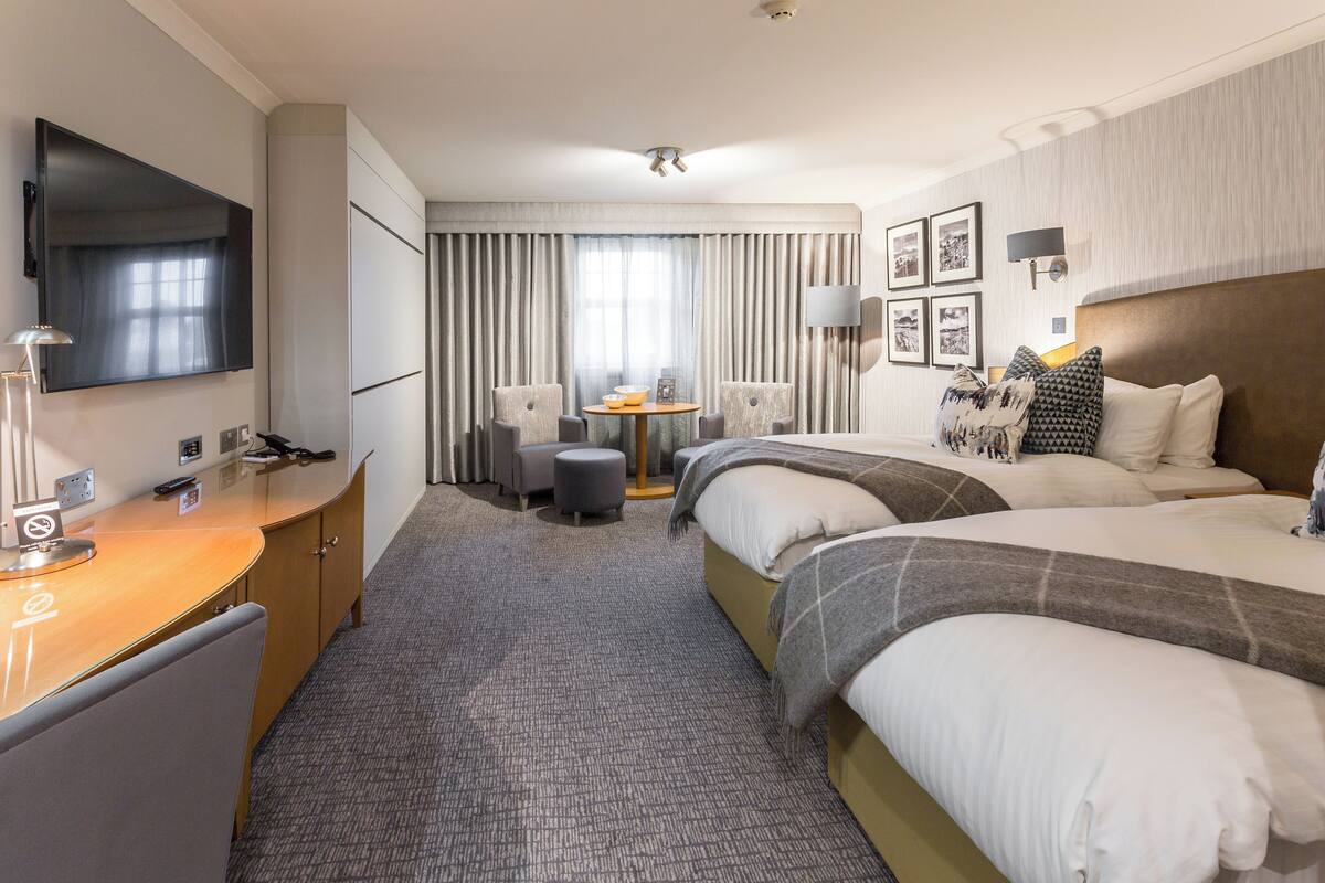 A hotel room with two beds, a desk with a chair, a TV, and a small table with chairs.