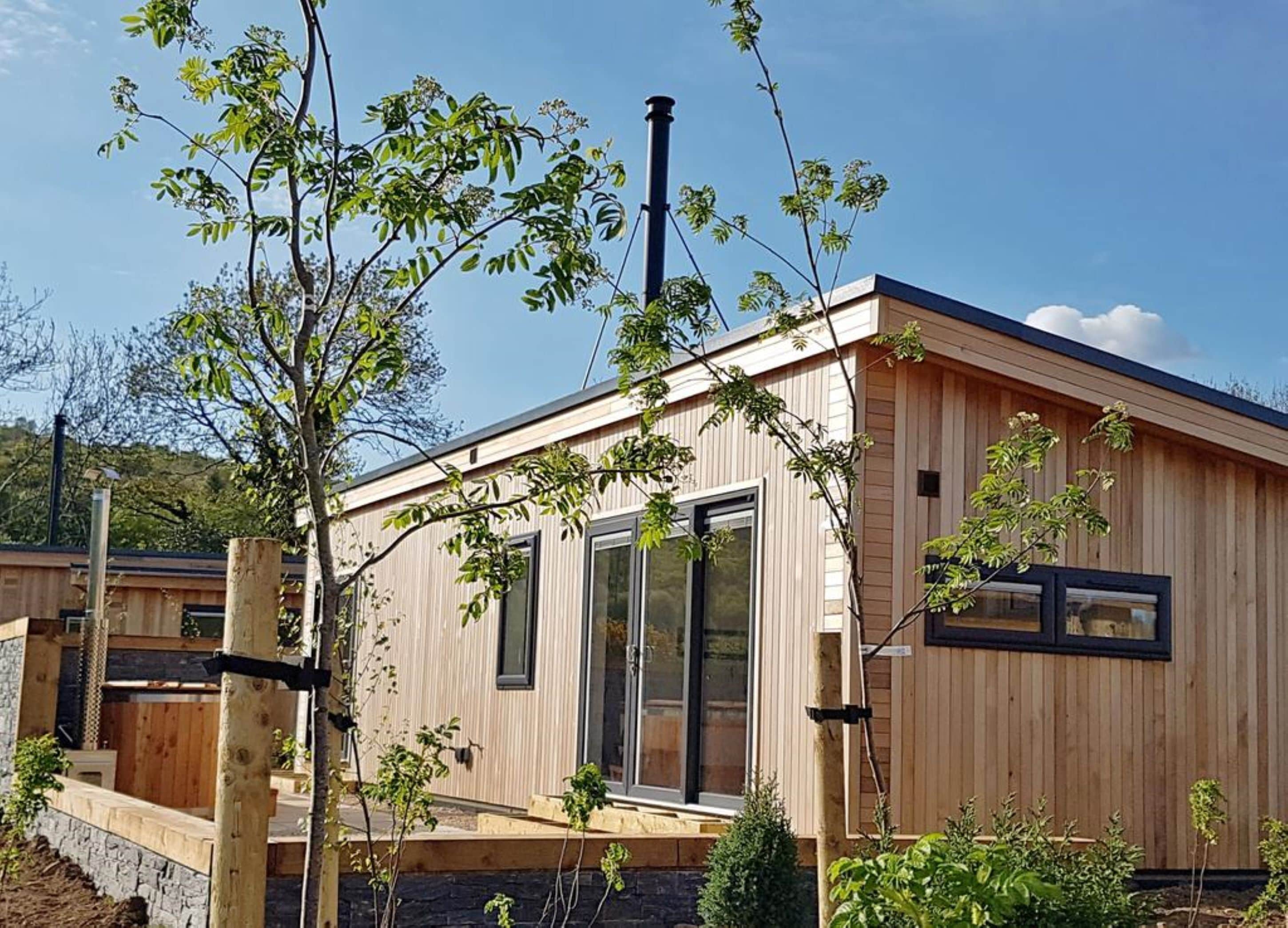 Una cabaña moderna de madera con ventanas grandes, cimientos de piedra y cielo despejado.