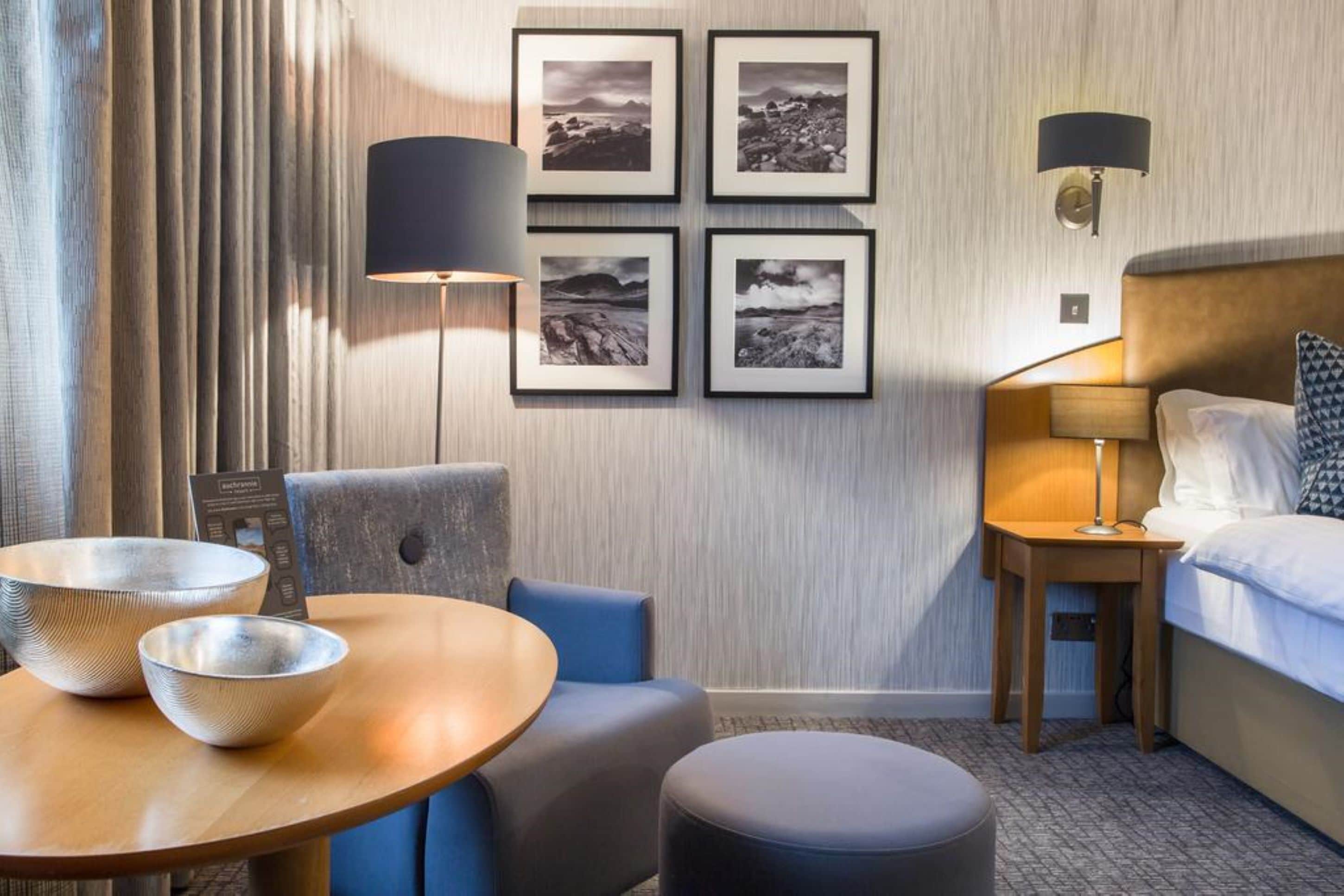 A hotel room with a bed, a round table with two bowls, a chair, a small ottoman, a wall-mounted lamp, and four framed pictures on the wall.
