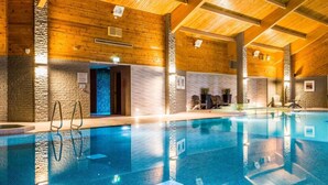 An indoor swimming pool with wooden beams and stone walls, surrounded by lounge chairs and a small seating area.