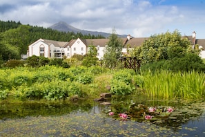 A large white building with multiple windows and a prominent entrance, surrounded by lush greenery and a pond with pink water lilies. - Auchrannie Resort (Isle of Arran)