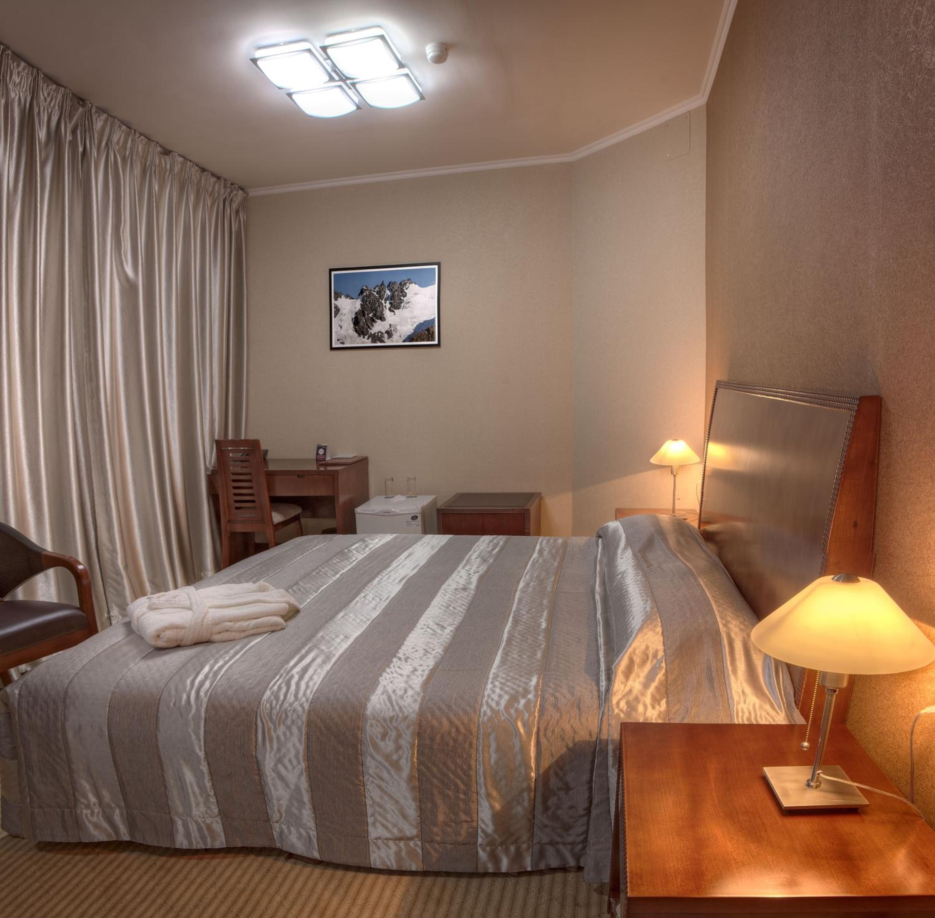 A hotel room with a bed, a desk, a chair, a lamp, and a picture on the wall.