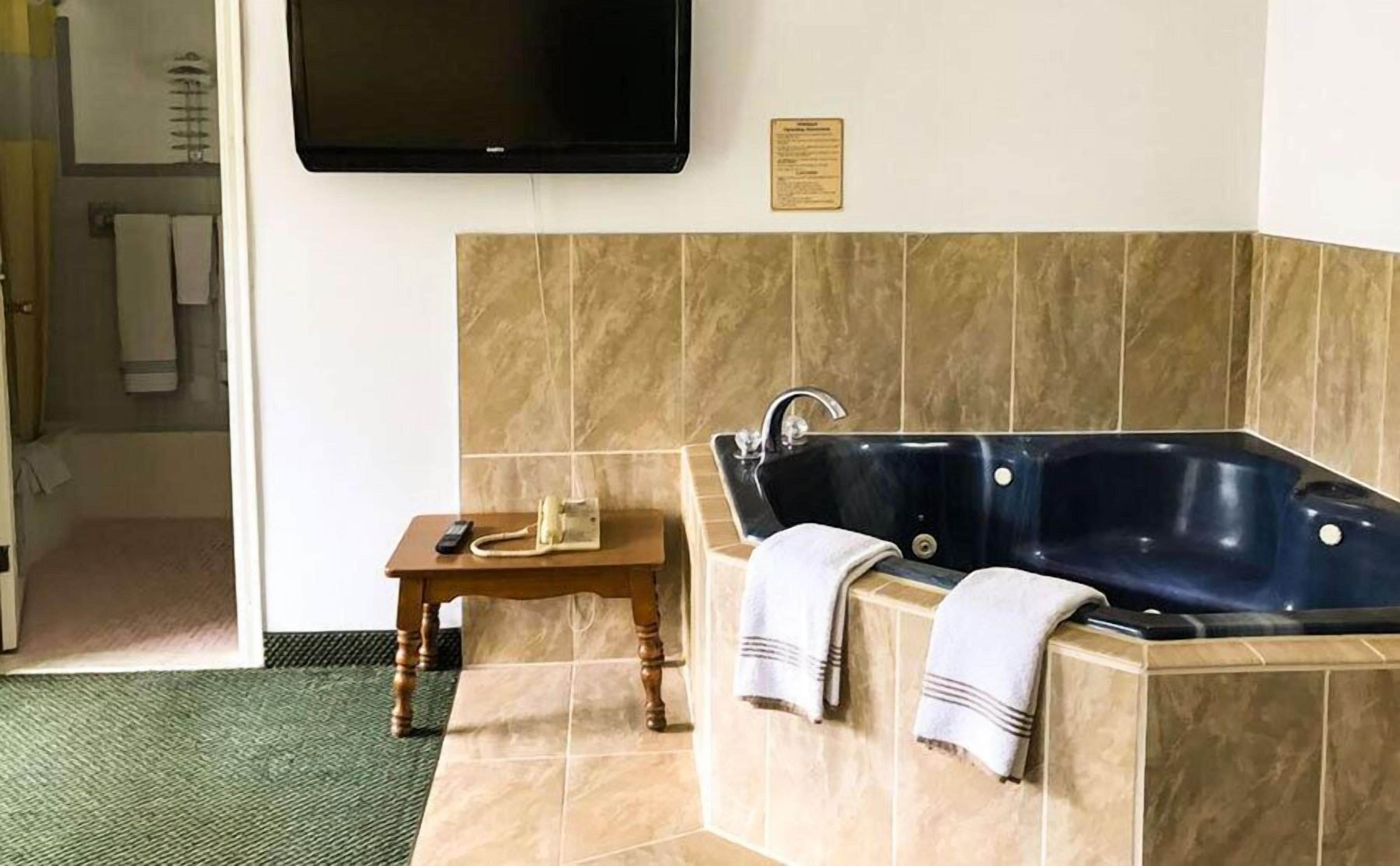 A bathroom with a large bathtub, a small wooden table with a telephone, and a wall-mounted TV.