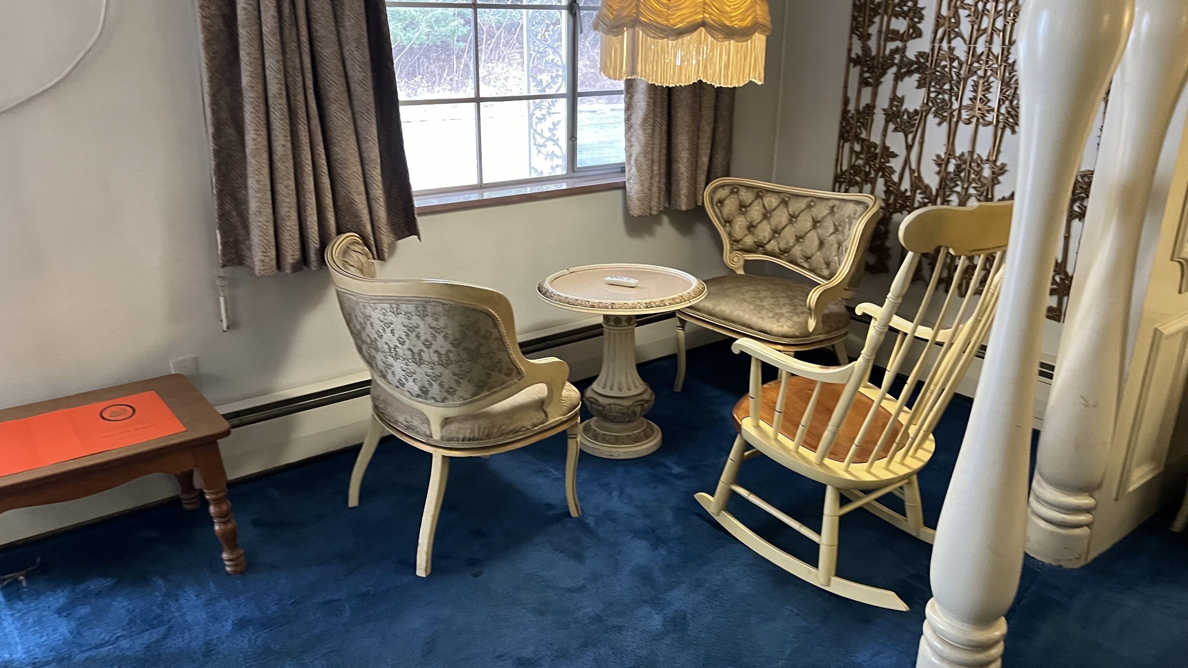 Une pièce avec un tapis bleu, une table ronde, deux fauteuils rembourrés, un fauteuil à bascule, une petite table sur laquelle se trouve un classeur rouge, et une fenêtre avec des rideaux.