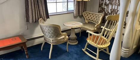 Une pièce avec un tapis bleu, une table ronde, deux fauteuils rembourrés, un fauteuil à bascule, une petite table sur laquelle se trouve un classeur rouge, et une fenêtre avec des rideaux.