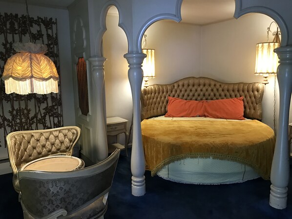 A bedroom with a tufted headboard, a four-poster bed, a chaise lounge, and a wall-mounted lamp.