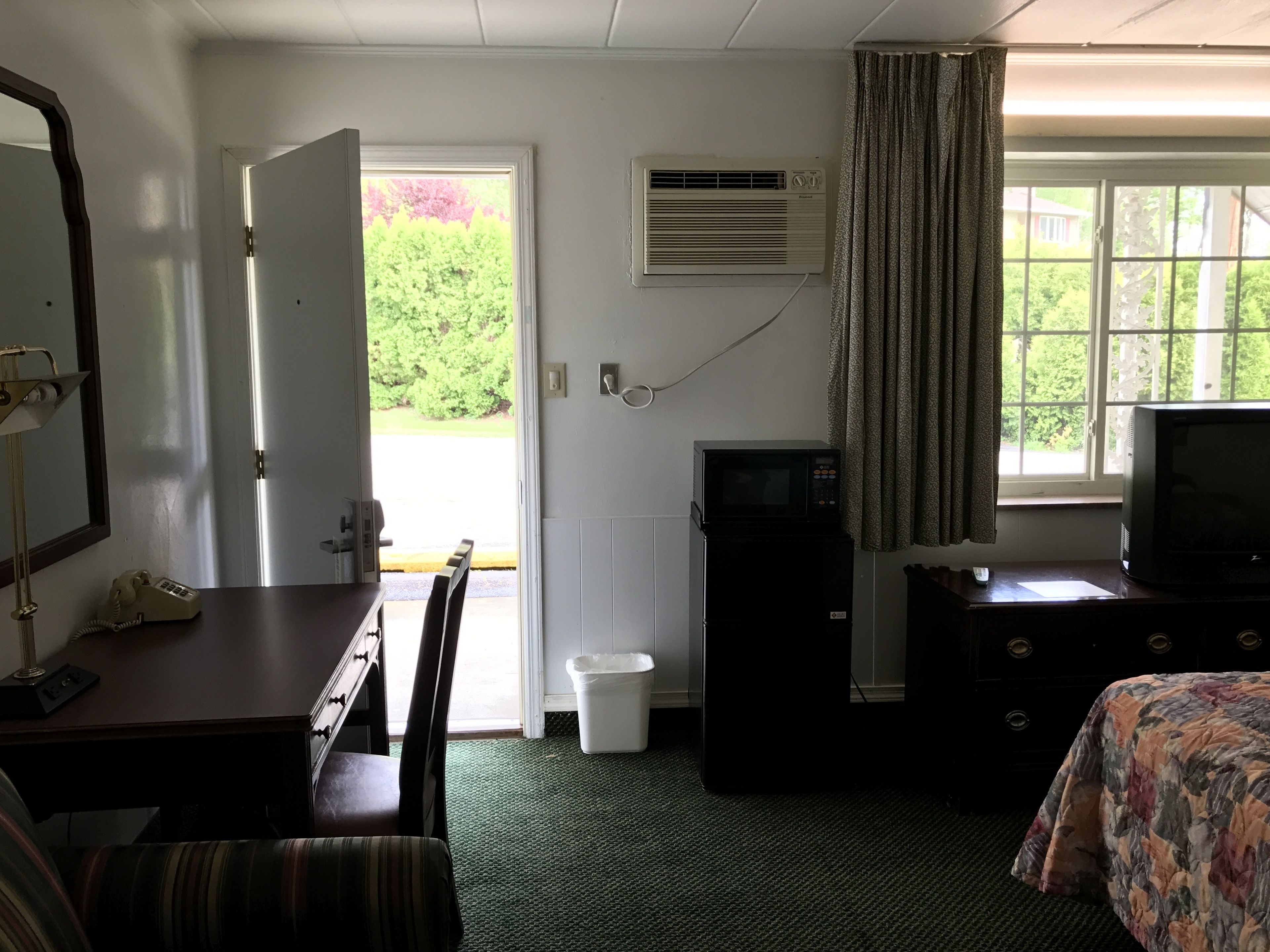Ein Hotelzimmer mit Schreibtisch, Stuhl, Bett, Fernseher und einem Fenster mit Vorhängen.