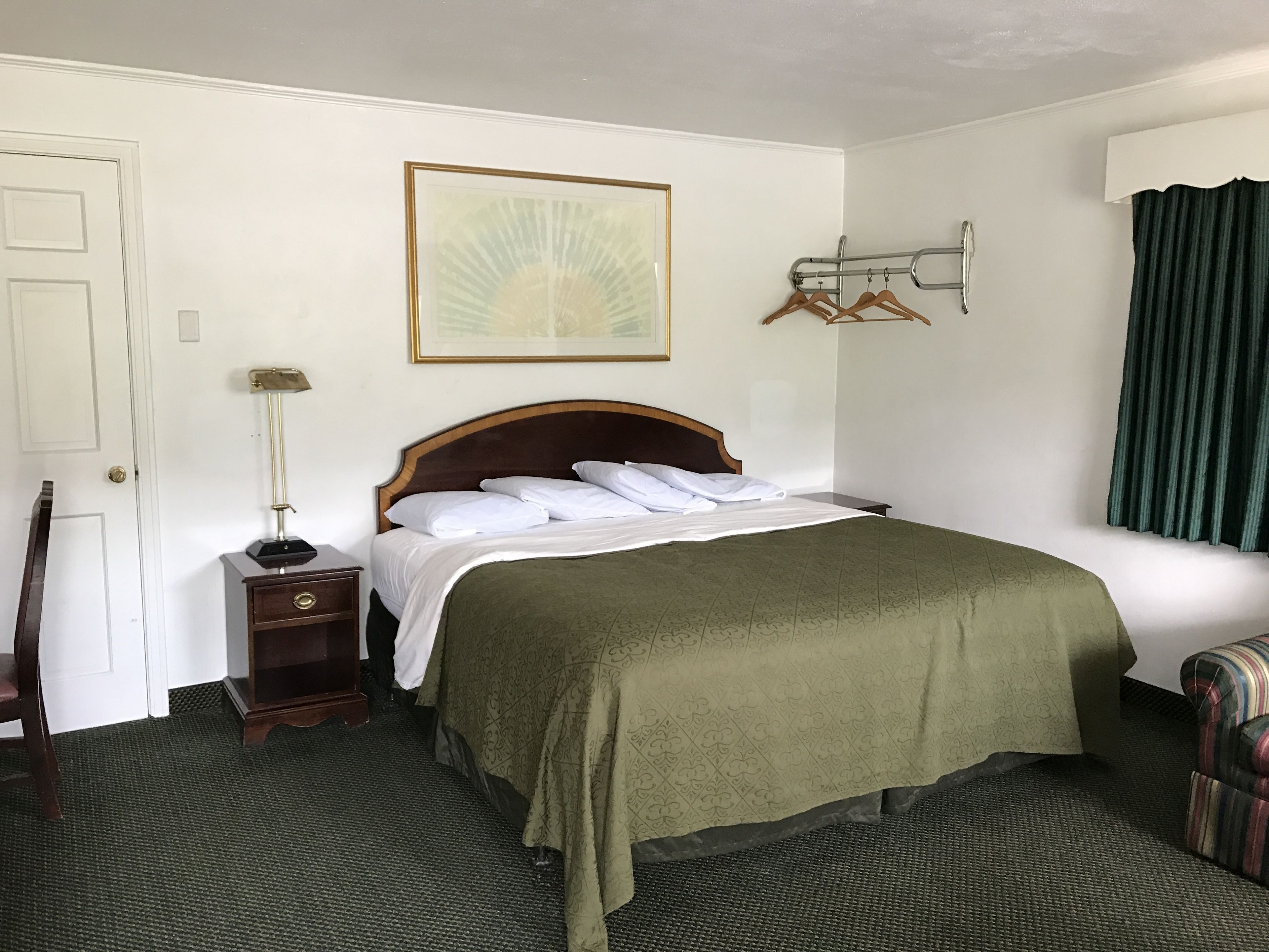 A neatly arranged hotel room with a bed, a bedside table, a lamp, a coat rack, and a window with curtains.