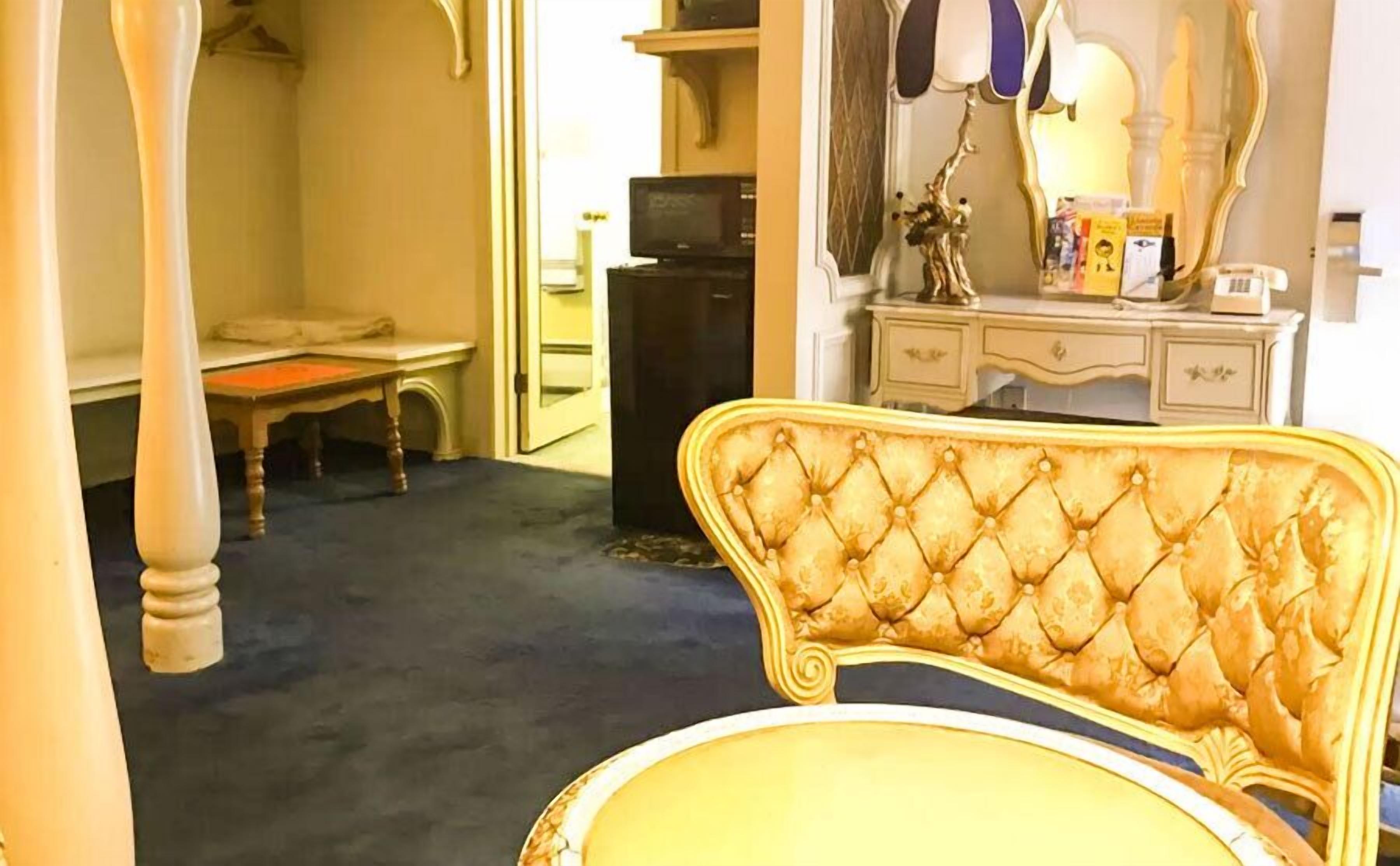 A room with a tufted headboard chair, a small table, a mirror, and a dresser.