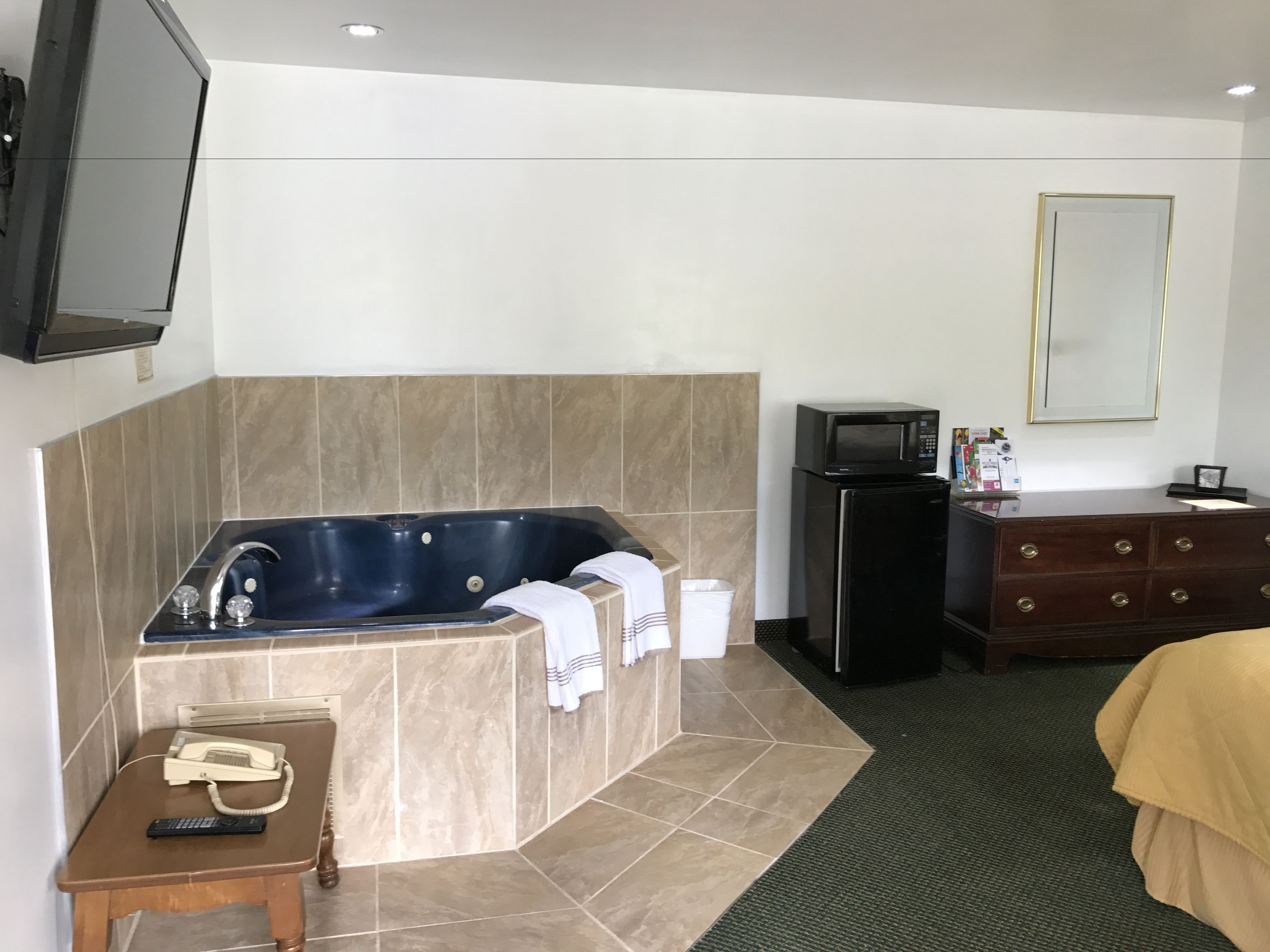 Ein Hotelzimmer mit einer großen Badewanne, einem kleinen Kühlschrank, einem hölzernen Schminktisch, einem Bett und einem an der Wand befestigten Fernseher.