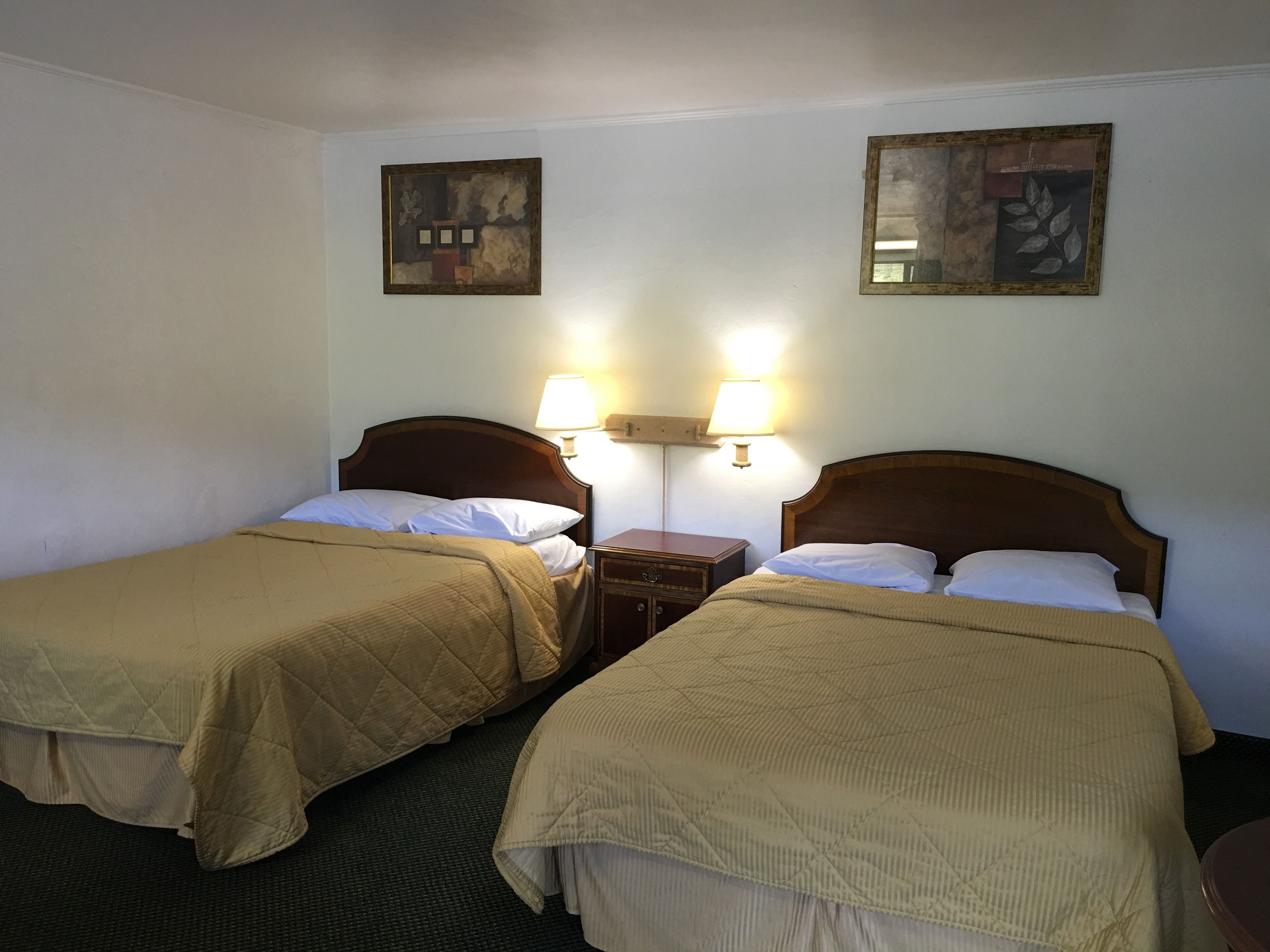 A hotel room with two single beds, each with a wooden headboard and a beige quilted bedspread. There are two wall-mounted lamps on either side of the beds, and two framed artworks above the beds.