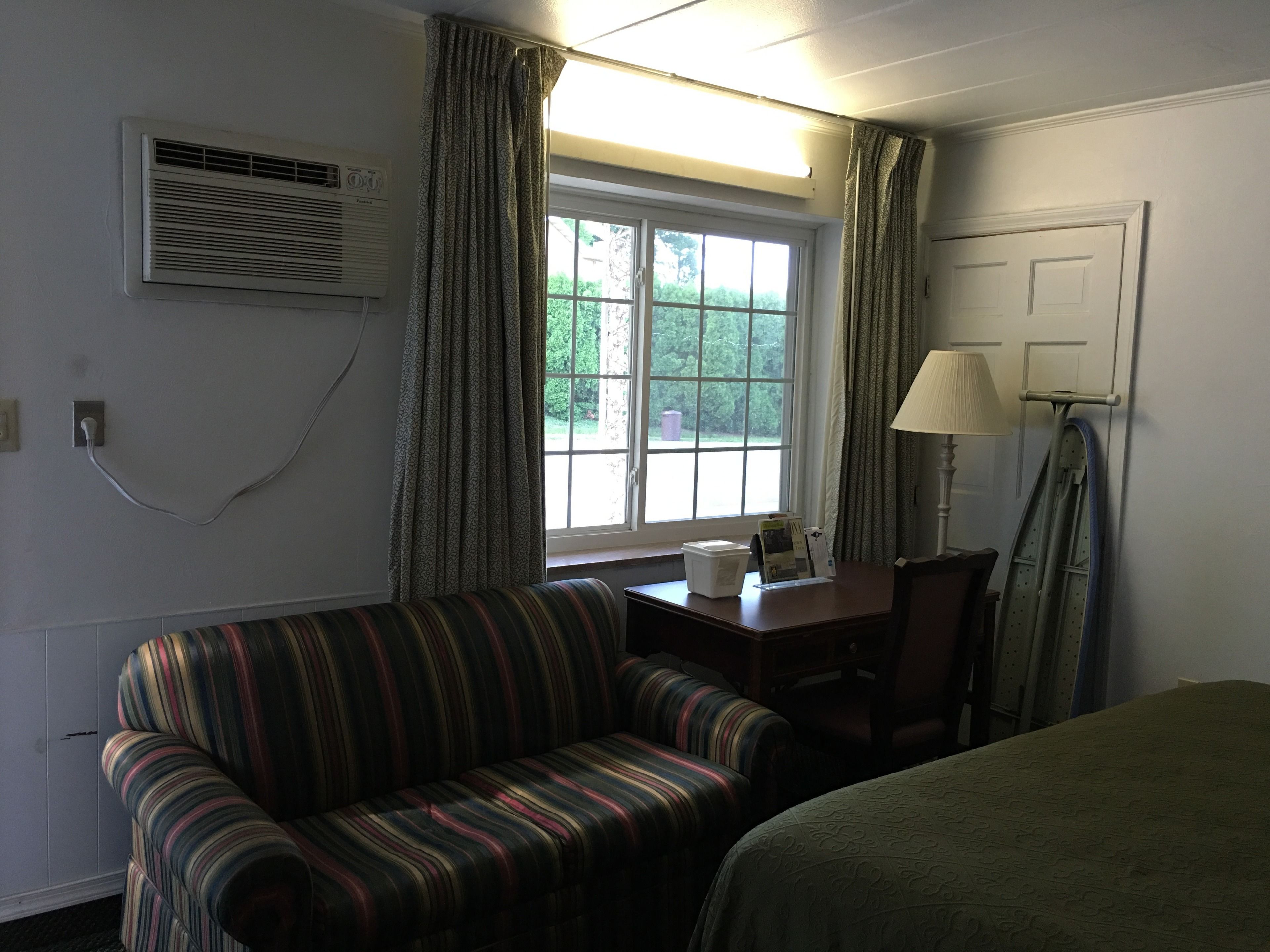 Een kamer met een gestreept sofa, een kleine tafel met een lamp, een raam met gordijnen en een airconditioner aan de muur.