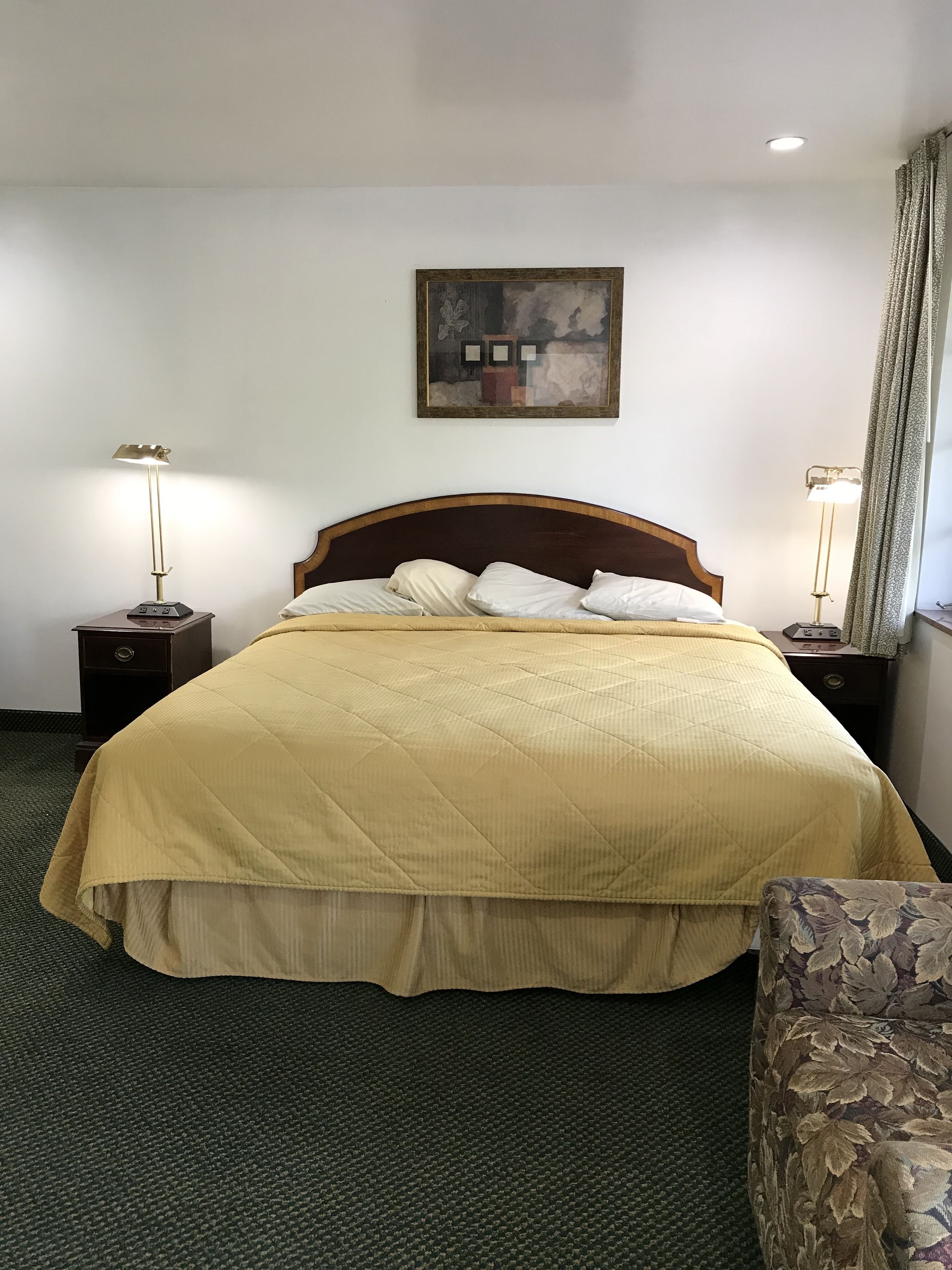 A hotel room with a large bed, two bedside tables, a nightstand, a chair, a painting on the wall, and a window with curtains.