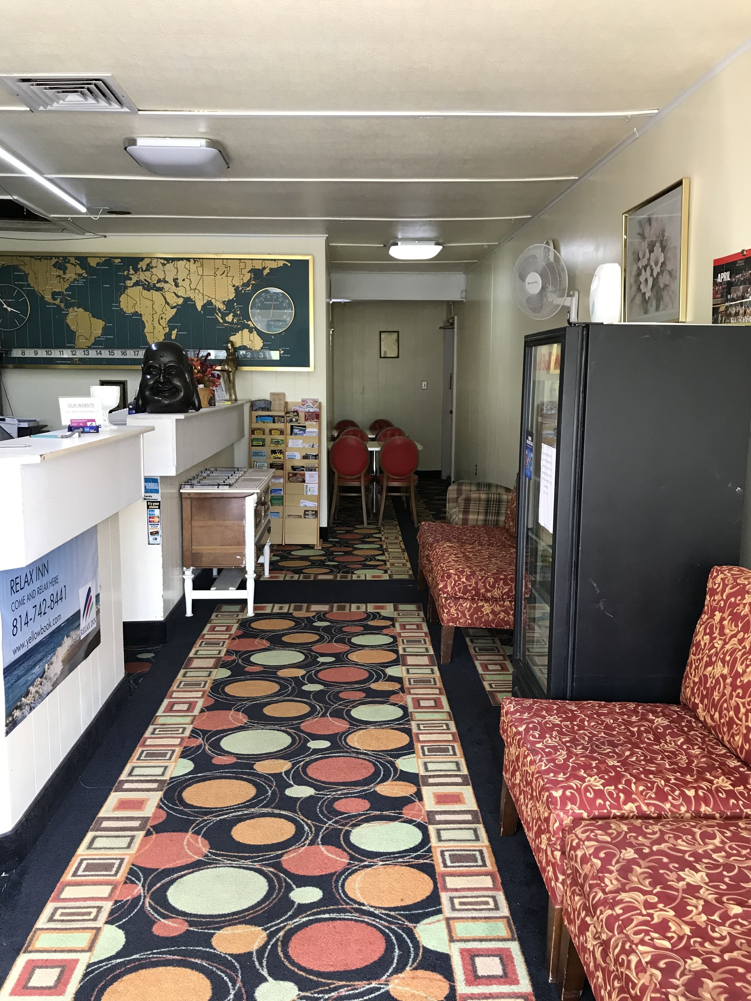 A hotel lobby with a reception desk, a world map, a clock, a refrigerator, and a patterned carpet.