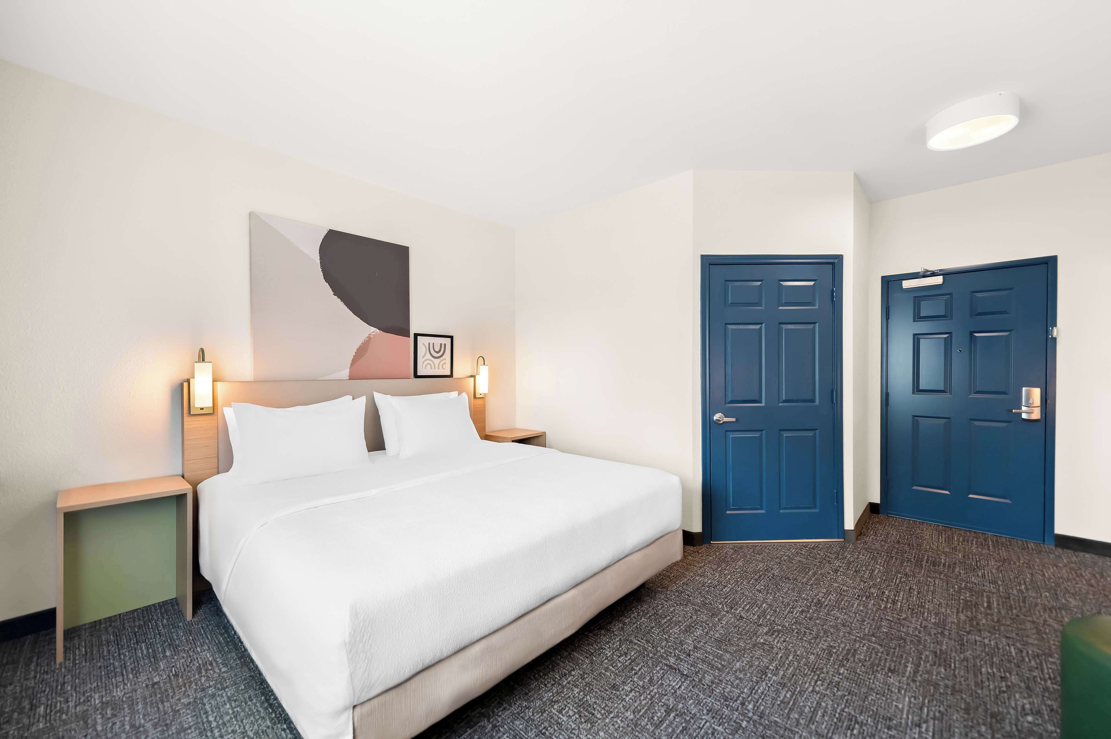 Ein Hotelzimmer mit einem großen Bett, zwei Nachttischen, einer blauen Tür und einem an der Wand hängenden Kunstwerk.